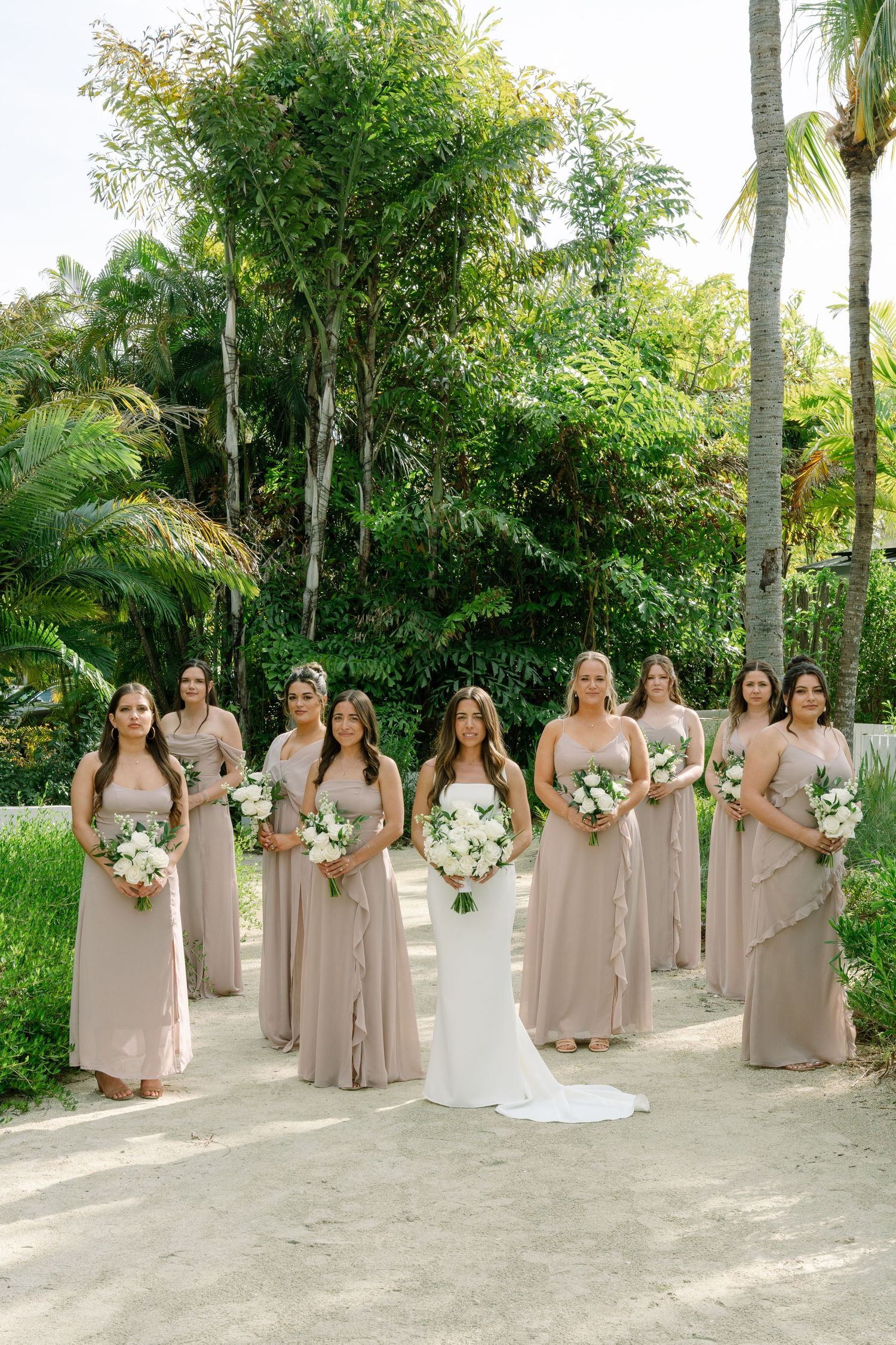 Bride with bridesmaids in taupe gowns holding white bouquets outdoors with lush tropical greenery.