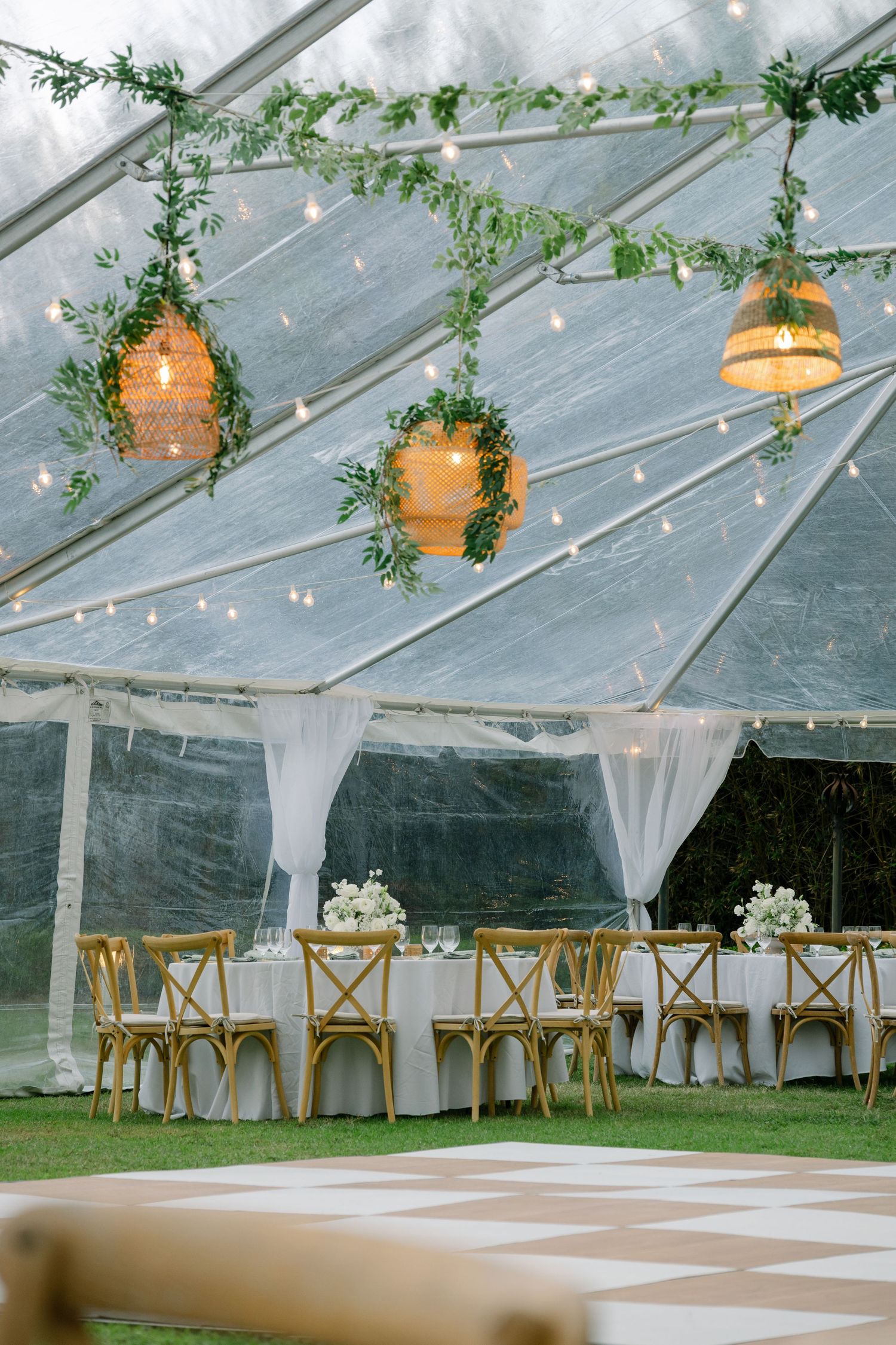 Elegant wedding reception under a clear tent with string lights, wicker pendant lights, greenery garlands, and gold cross-back chairs.
