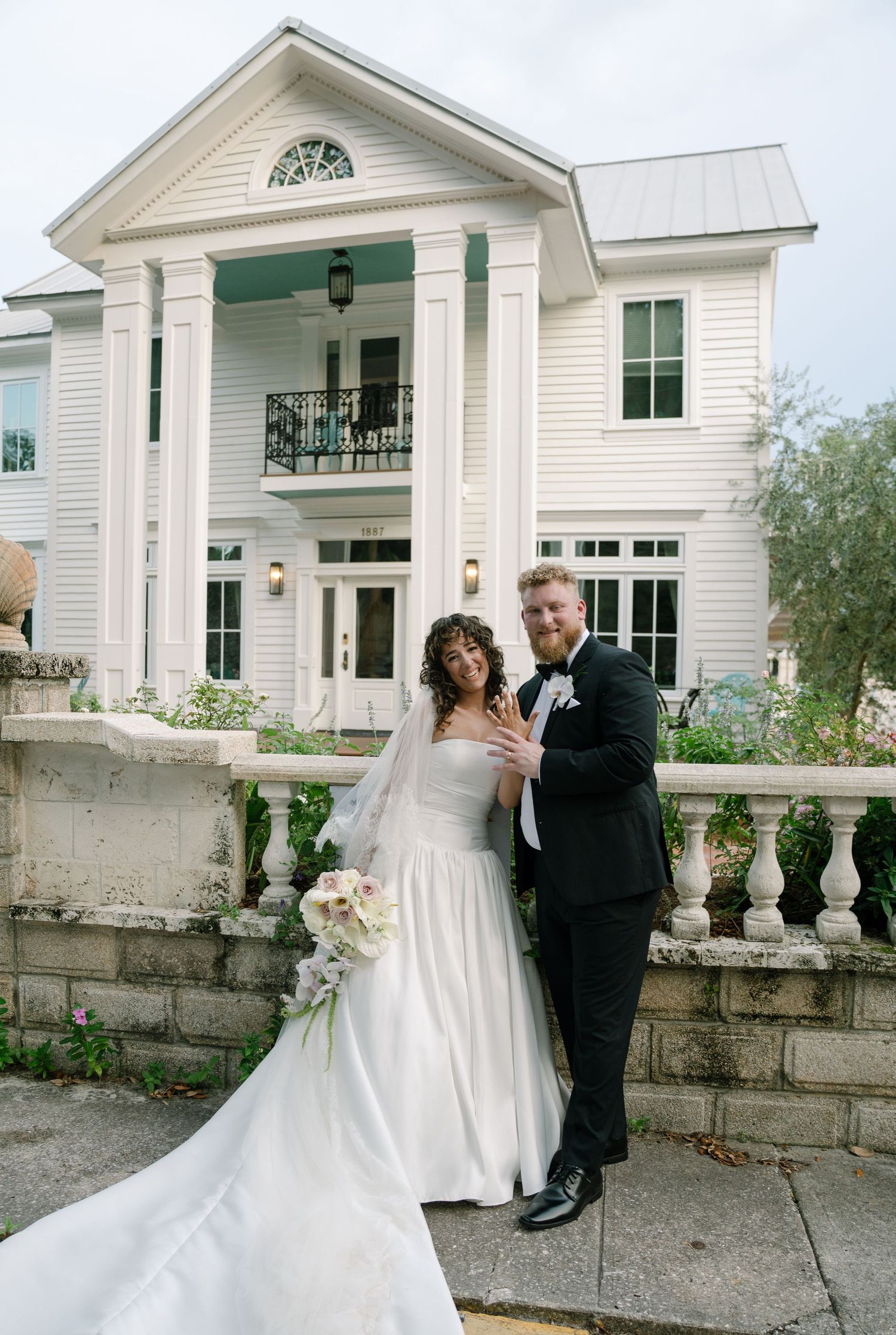 Wedding couple posing in front of a white colonial style building with stone wall.