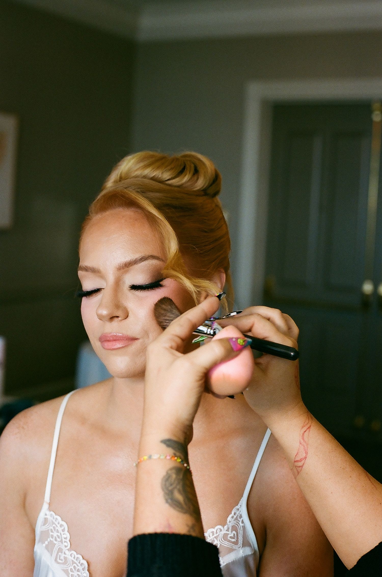A makeup artist applies mascara to a person wearing a white lace top in a series of close-up shots.