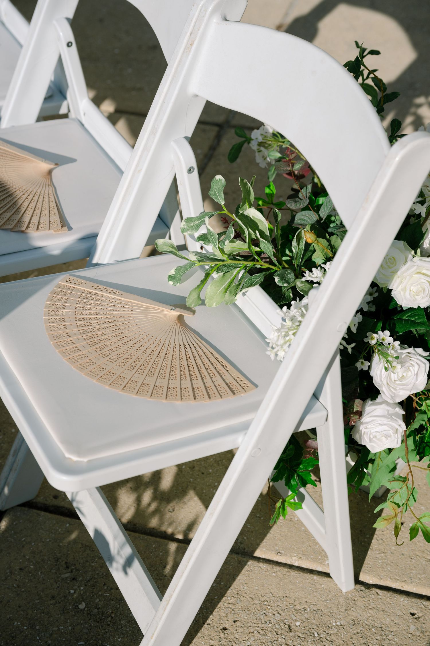 White folding chairs decorated with beige hand fans and floral arrangements for an outdoor wedding ceremony.