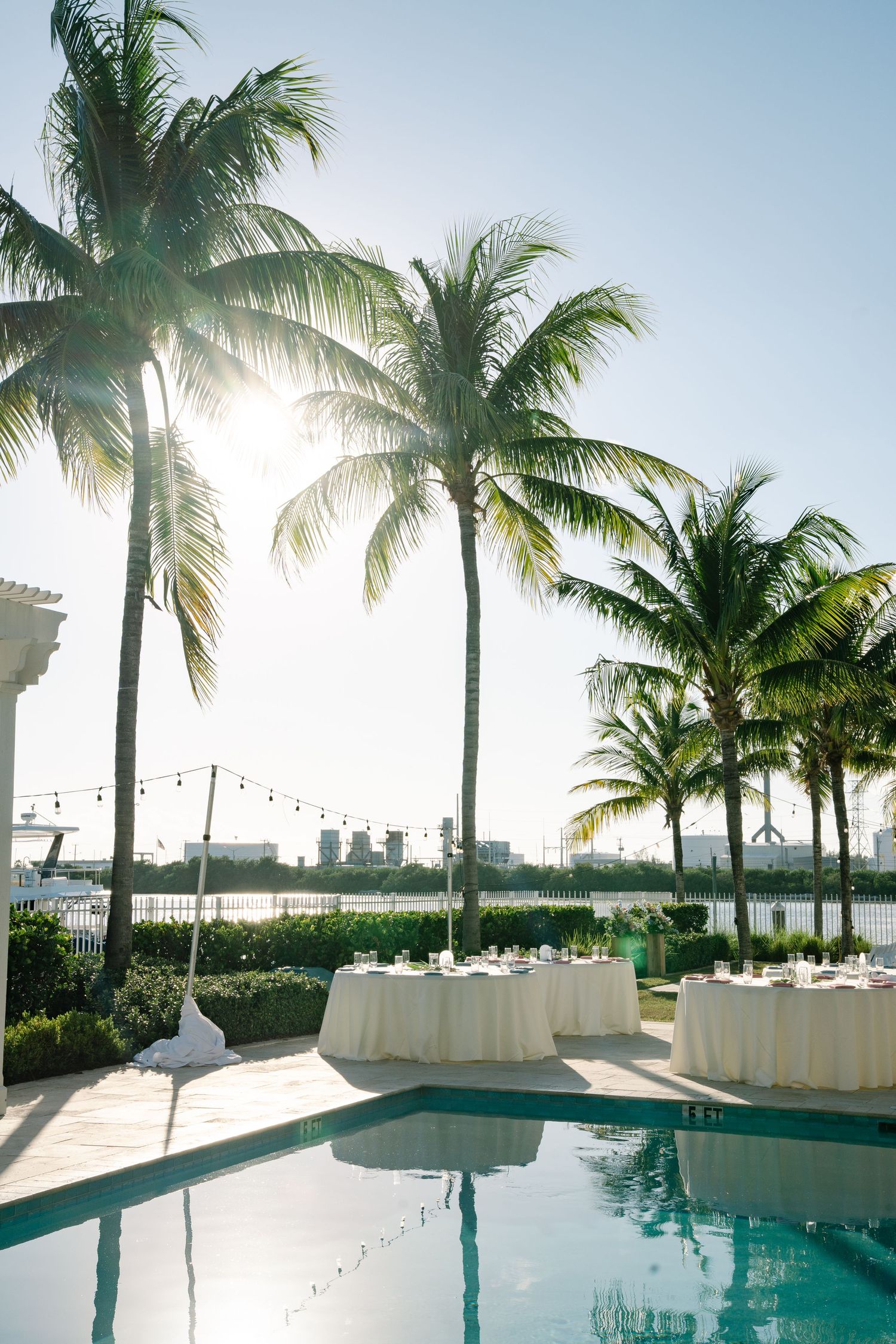 Poolside wedding reception setup with palm trees and string lights at sunset.