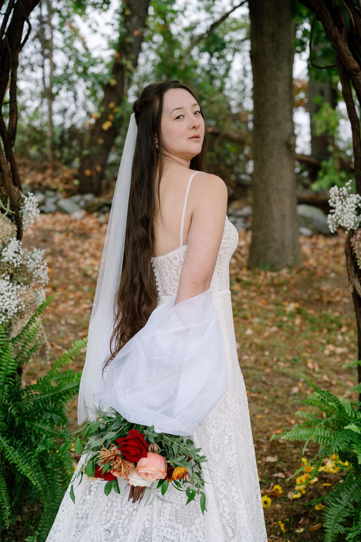 A bride in a white lace dress holds a colorful bouquet while standing in a wooded outdoor setting.