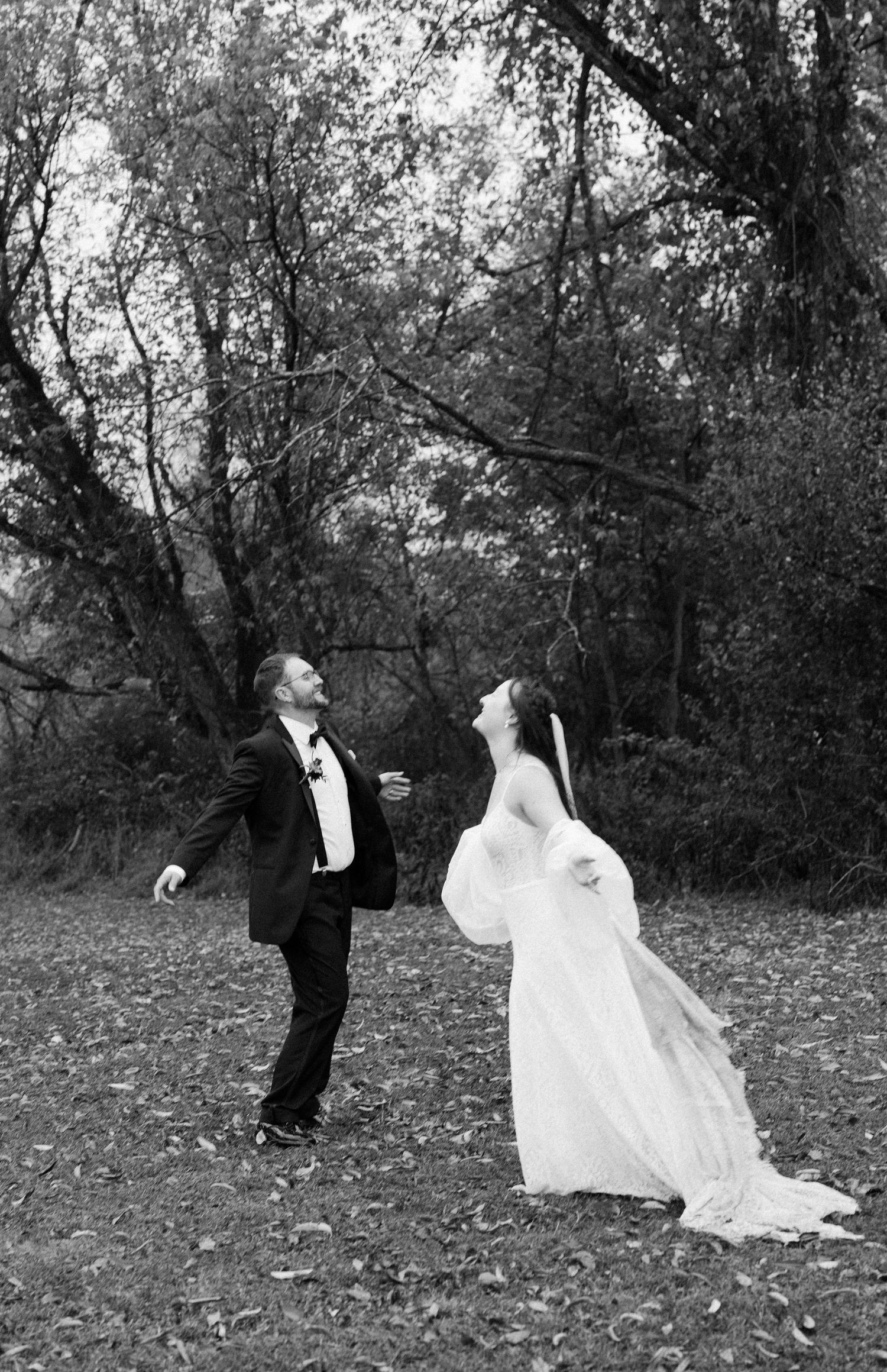 Wedding couple twirling and dancing joyfully among autumn trees in a black and white photograph.