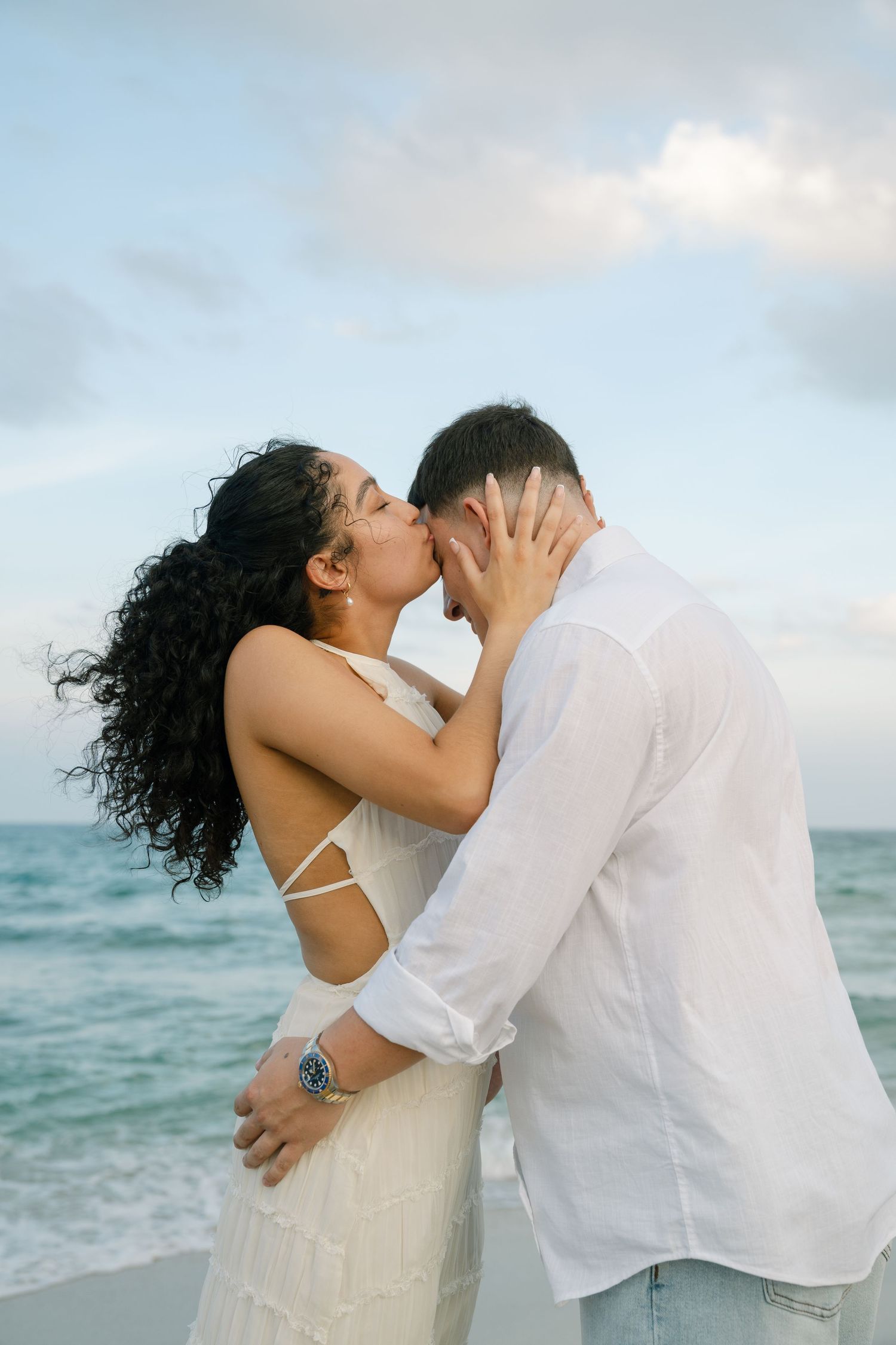 A couple embraces and kisses passionately on a scenic beach with turquoise waters behind them.