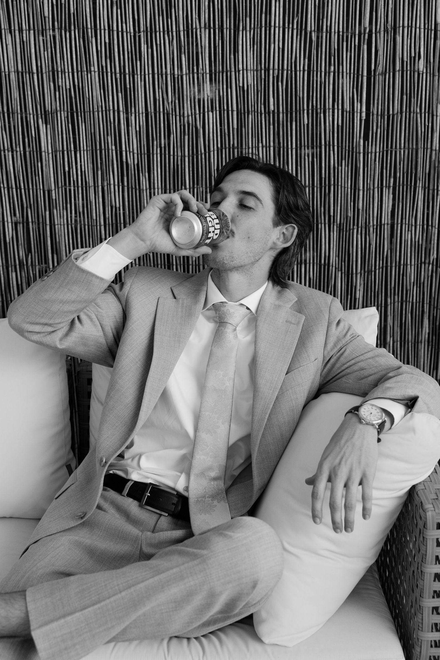 A man in a suit sits on a chair, drinking from a glass, against a backdrop of bamboo sticks.