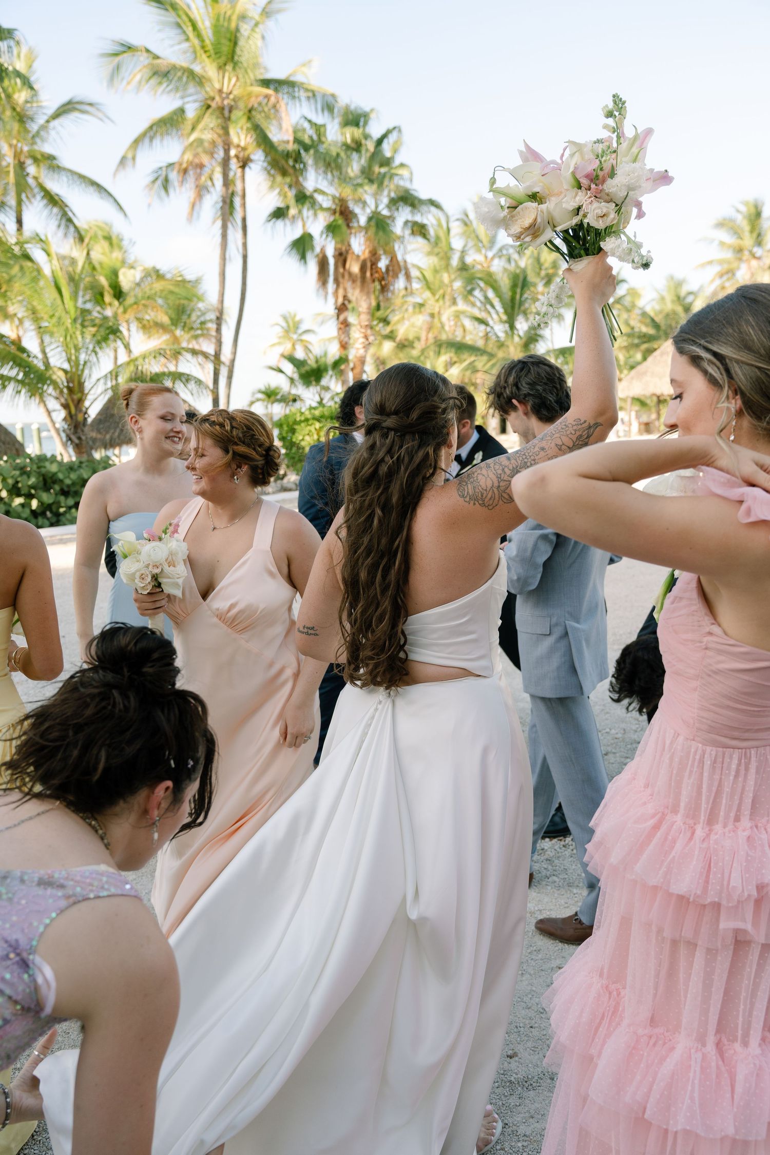 Bride and bridesmaids in pink and white dresses celebrate on a tropical beach with palm trees, holding floral bouquets.