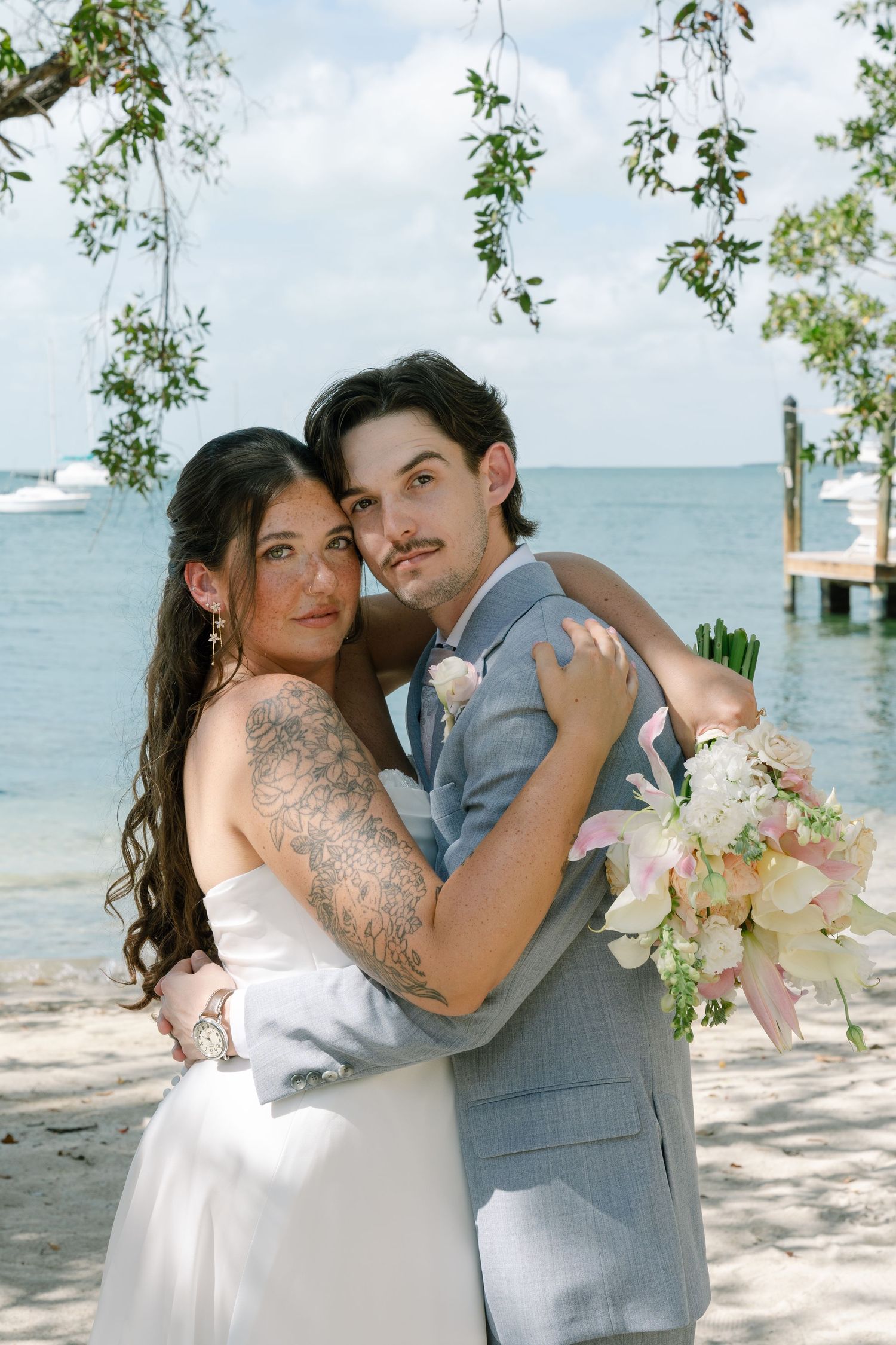 A wedding photo of a couple embracing on a beach with trees and water in the background.