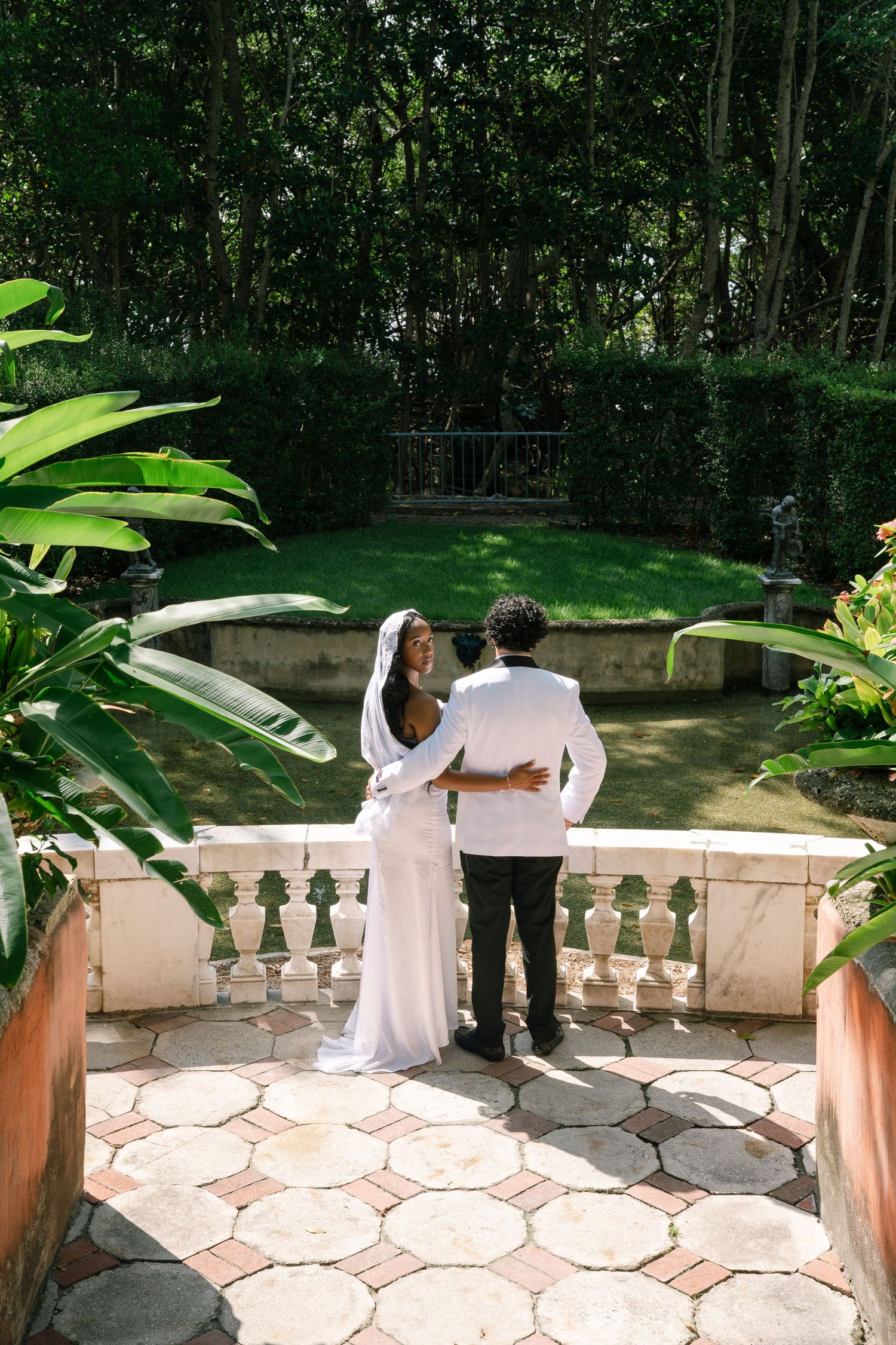 A couple stands on a stone terrace overlooking a lush garden with tropical plants and ornate stone balustrades.