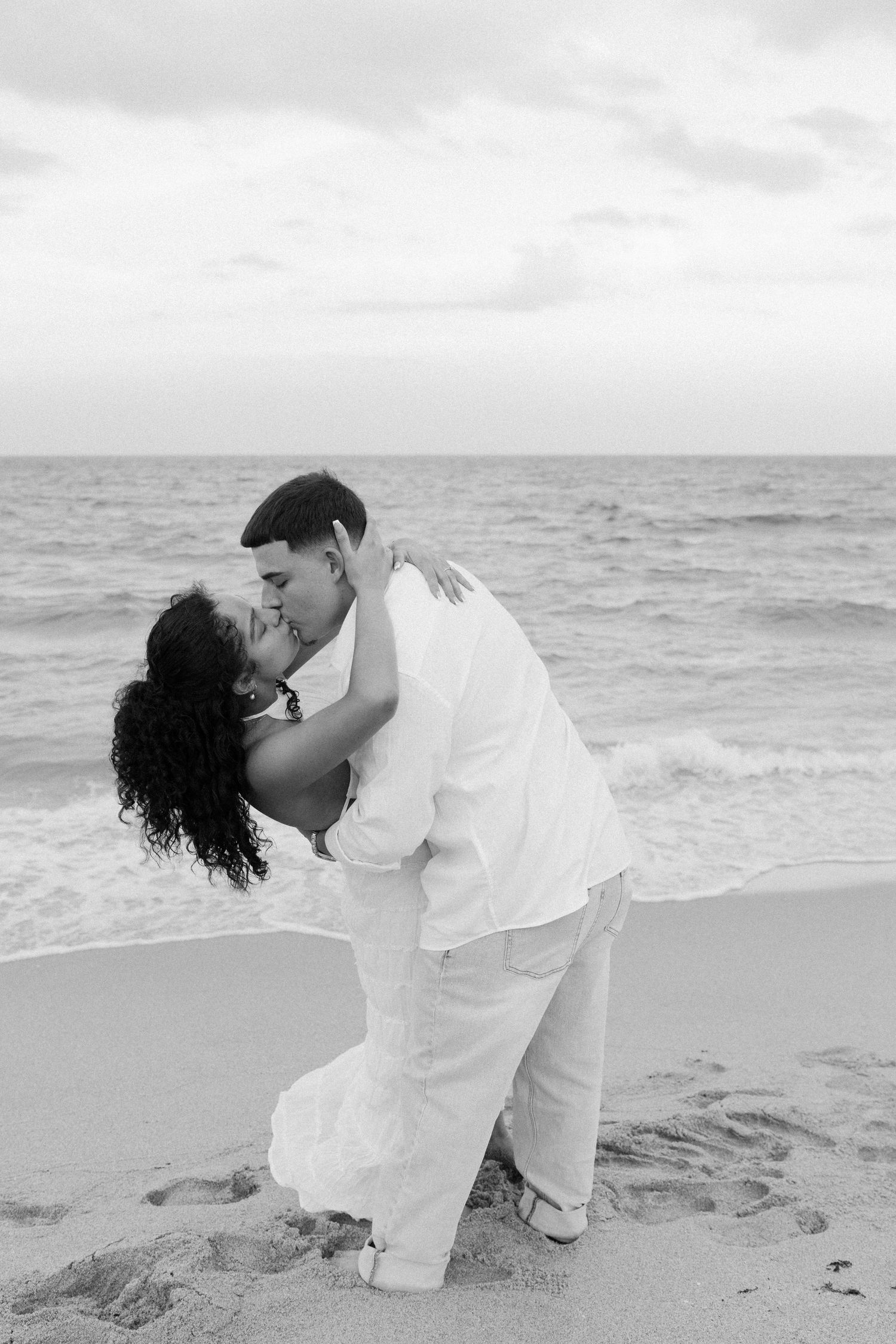 A couple in white attire share a romantic dip pose on a sandy beach with ocean waves in the background.