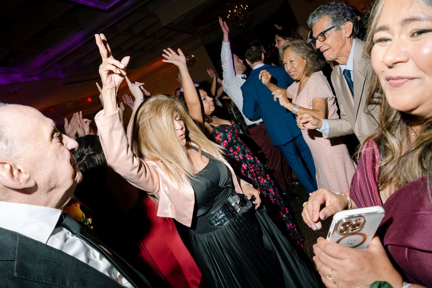 Wedding reception guests dancing and celebrating on a dimly lit dance floor with purple lighting.