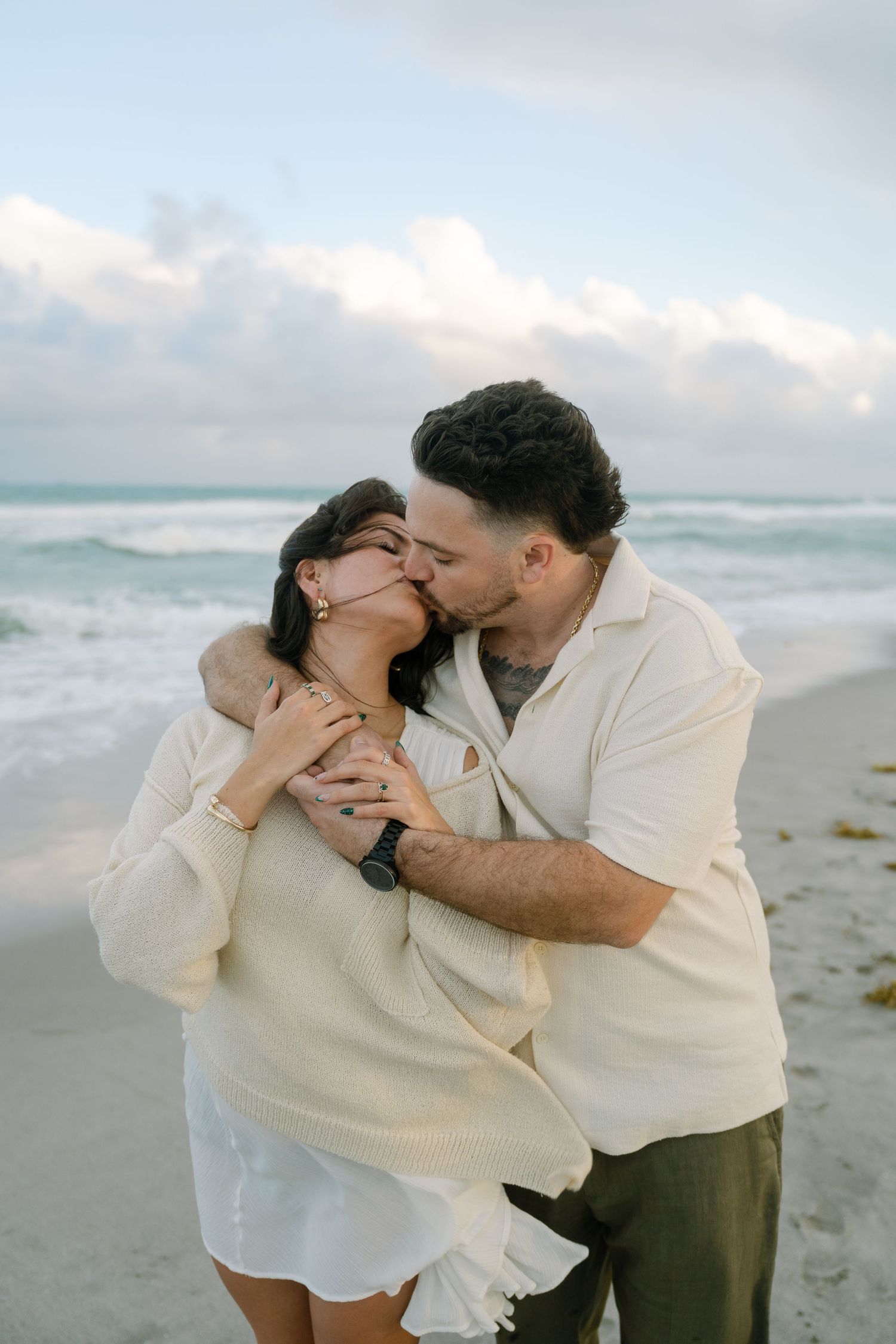 A romantic couple shares tender moments together on a cloudy beach, wearing matching cream colored outfits.