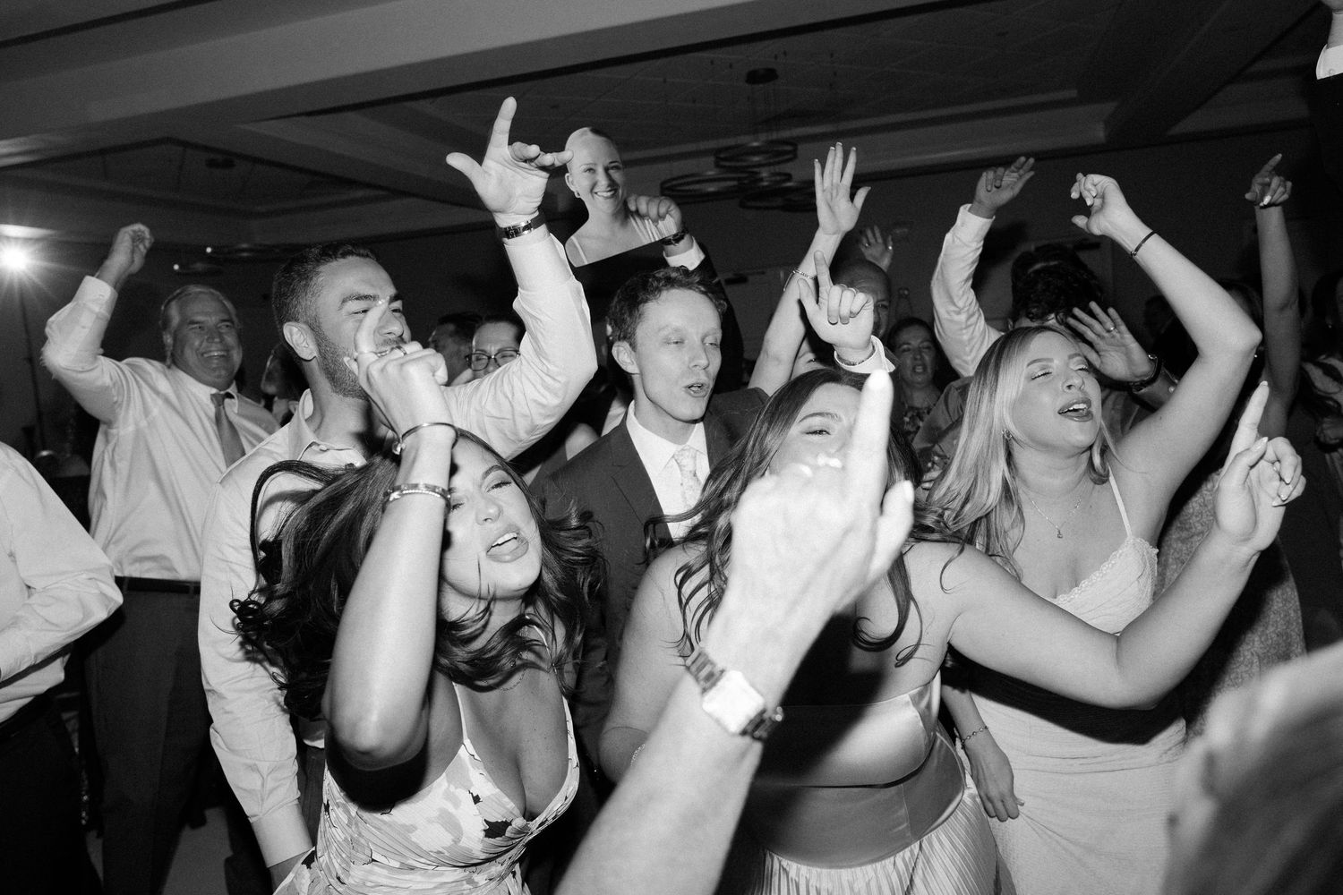 A group of people dance and celebrate with raised hands at a wedding party in black and white photography.