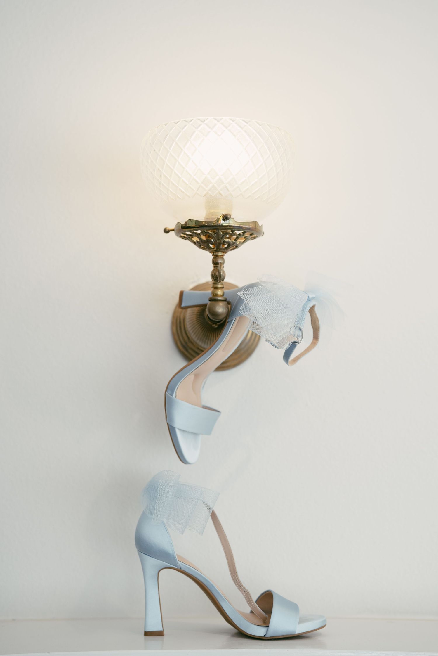 Elegant light blue suede heeled sandals with ankle straps displayed beneath an ornate vintage brass wall sconce.