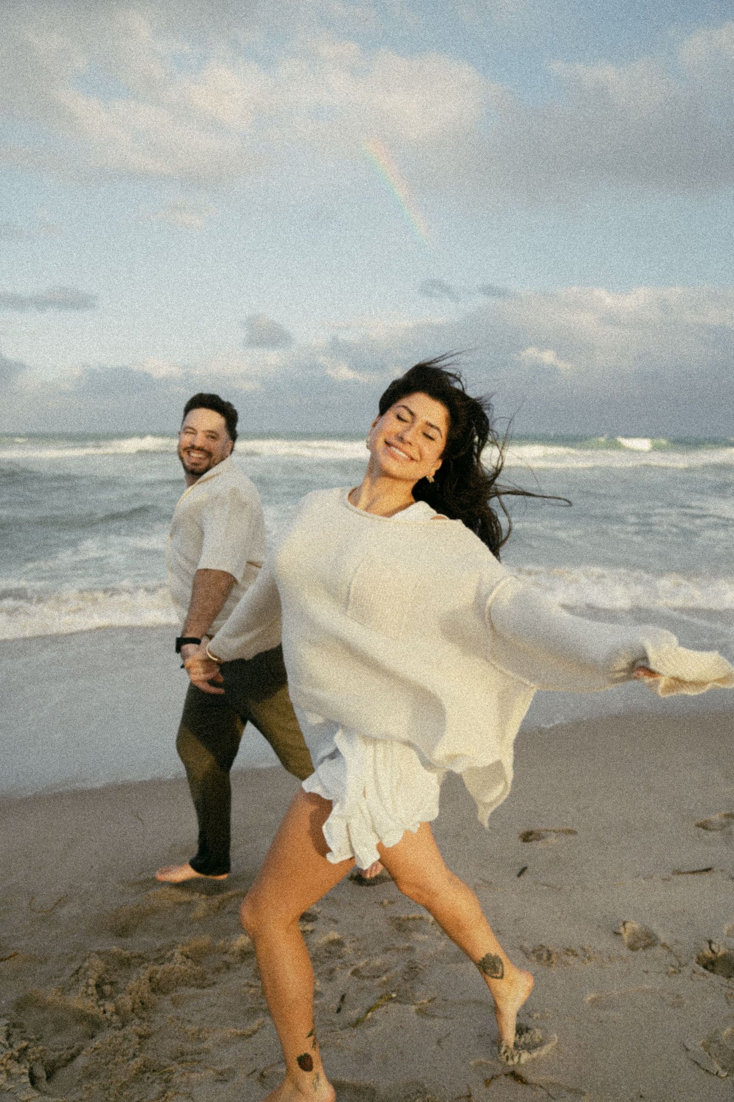 Couple holding hands and playing joyfully together on a windy beach shore as waves crash behind them.