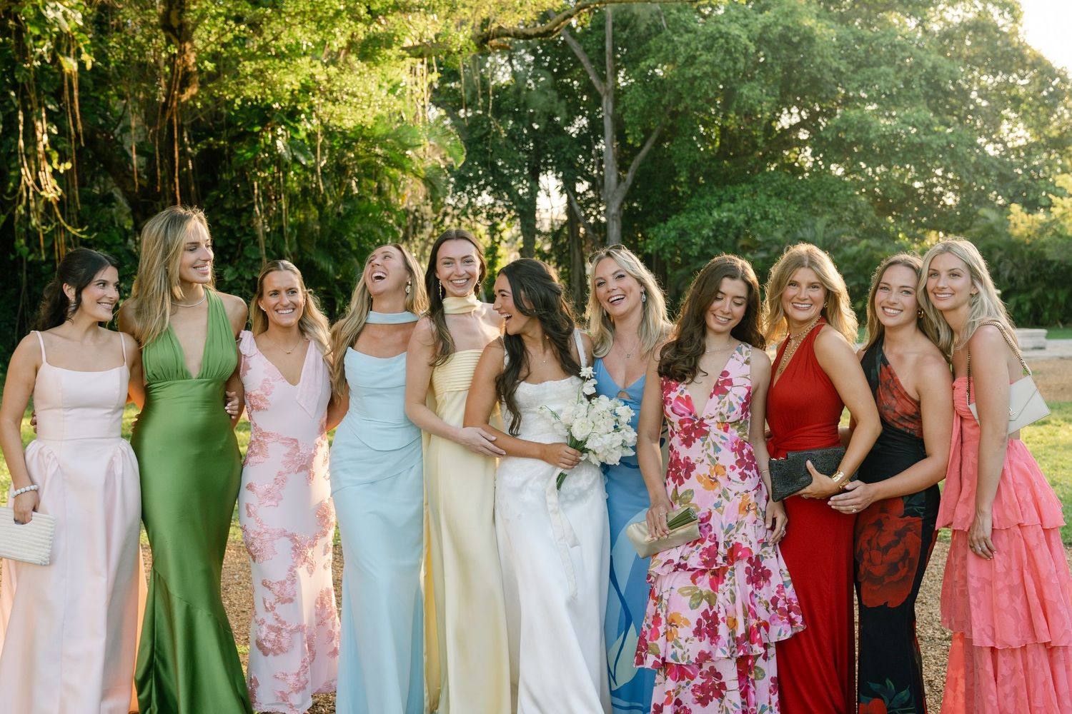 Bride poses with bridesmaids in colorful mismatched gowns laughing in lush garden setting.