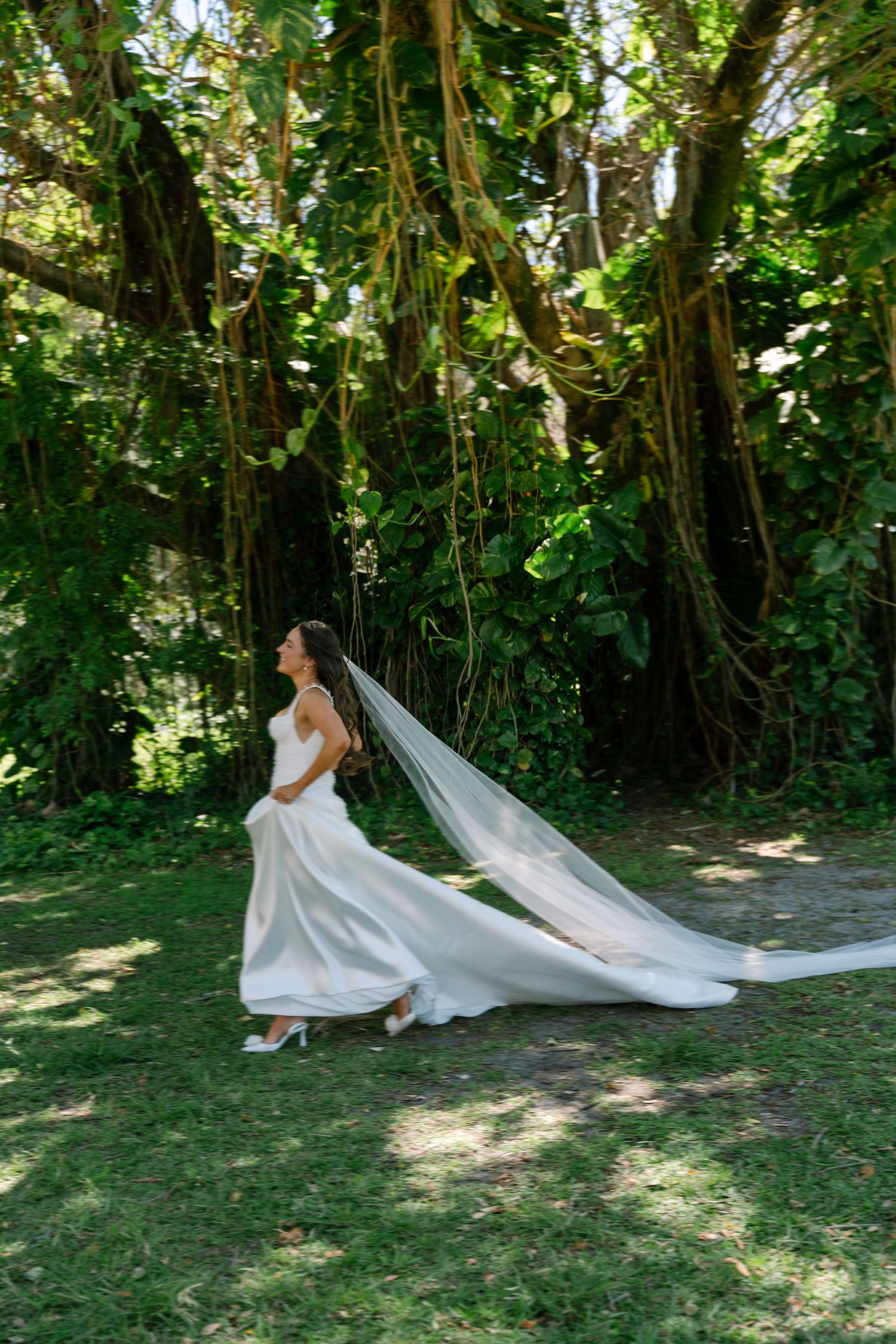 Bride in strapless white gown with long veil walks through lush tropical garden on wedding day.