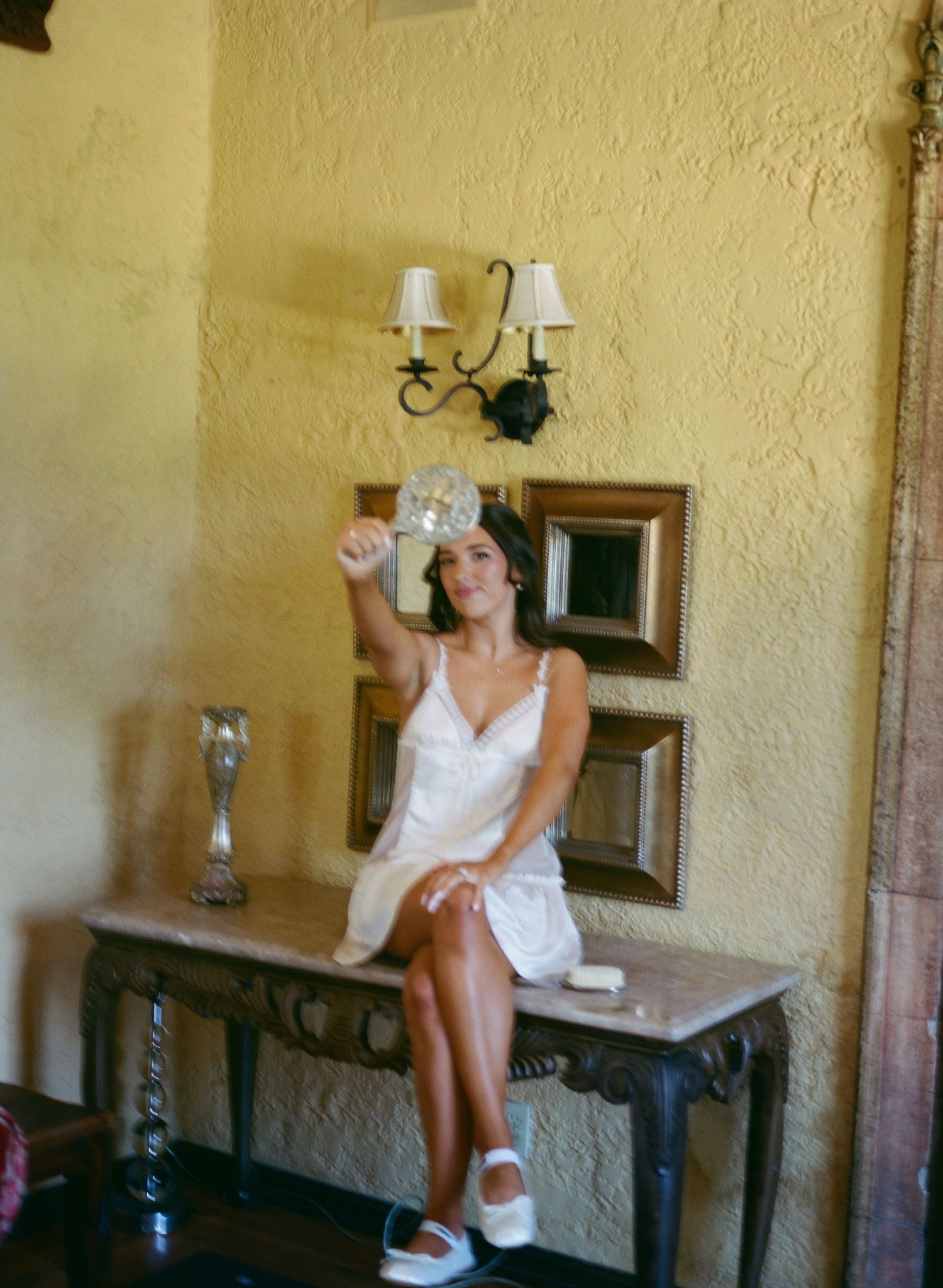 Bride in white slip dress sits on marble console table holding a decorative fan indoors.