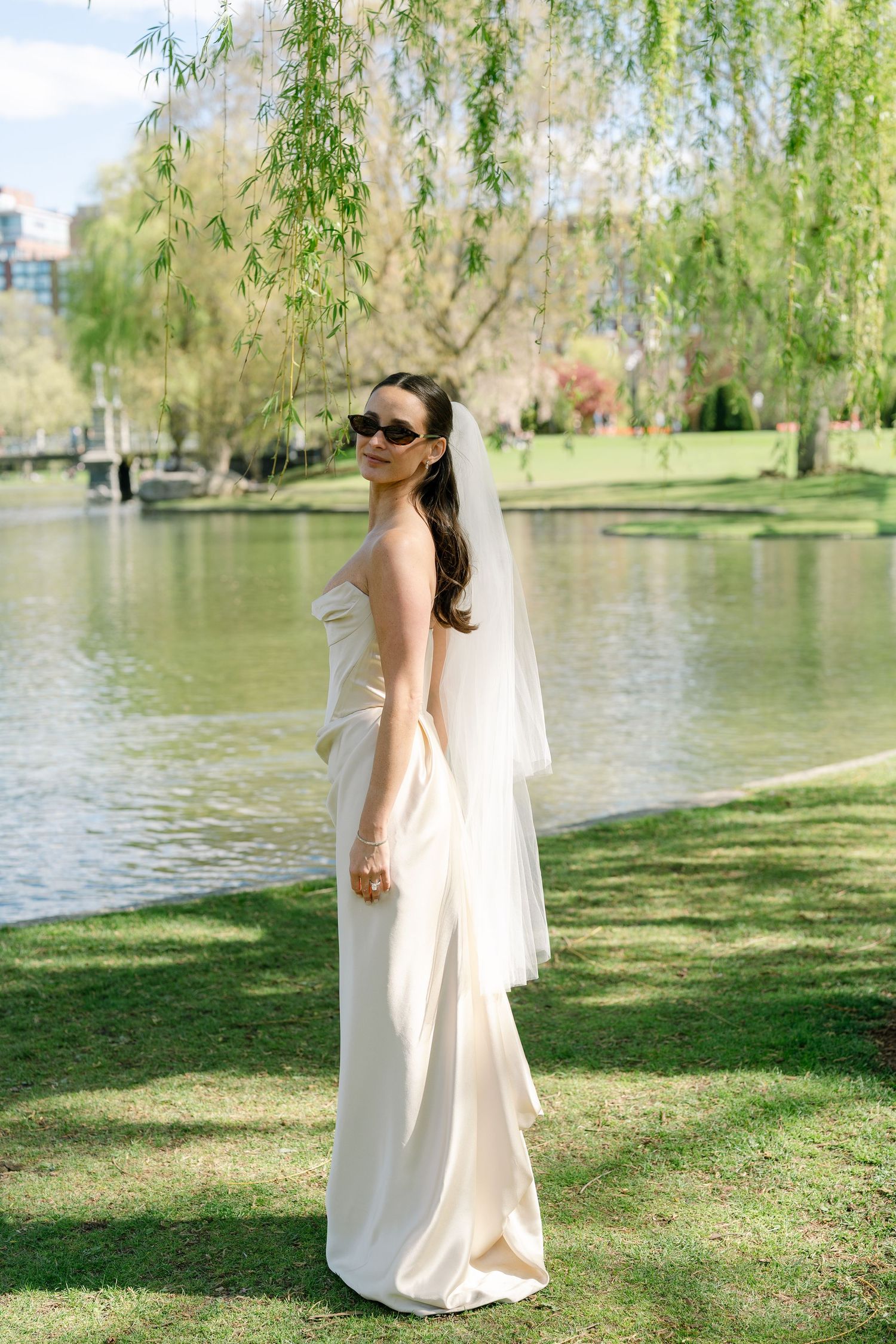 Elegant bride in white strapless gown and veil wearing sunglasses, posing by a serene pond in a lush green park.