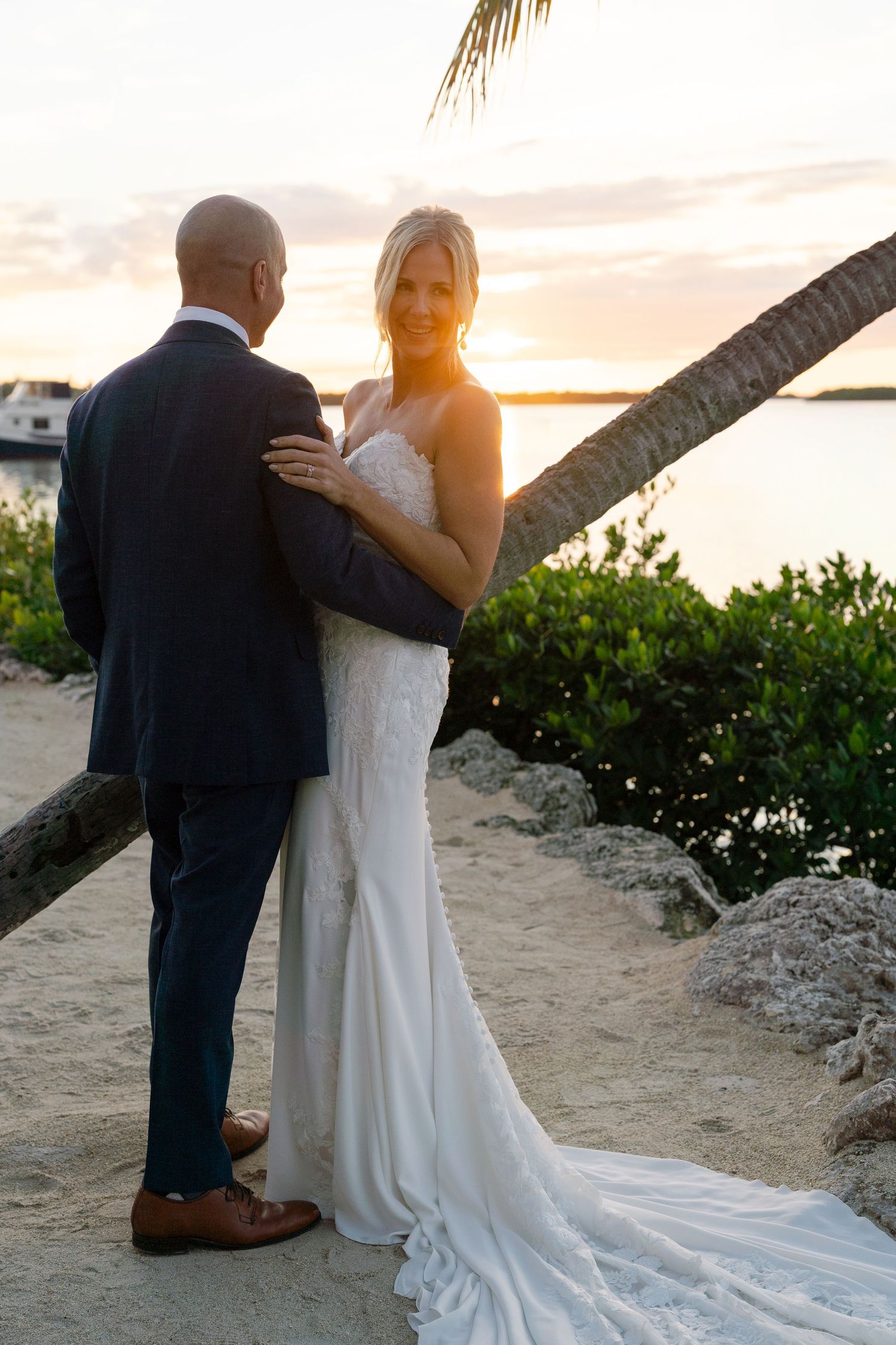 Newlyweds share an intimate moment at sunset on a tropical beach with palm trees.