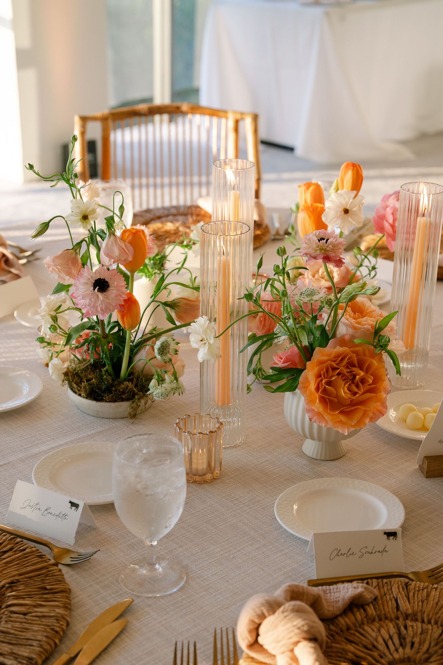 Wedding table setting with amber glass candlesticks, orange and pink floral arrangements, and place cards on woven chargers.