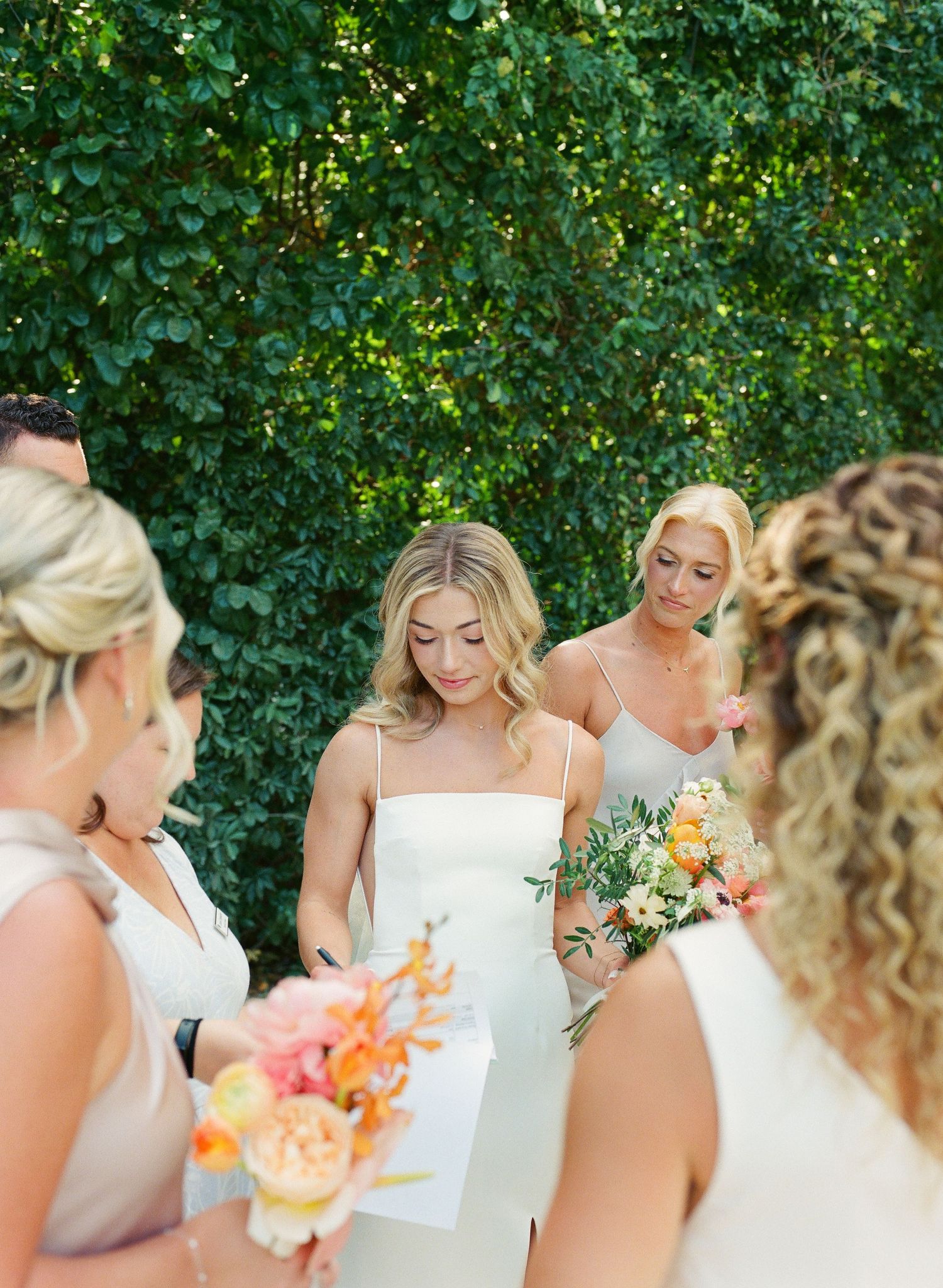 Wedding party gathering outdoors with floral bouquets against green hedge backdrop.