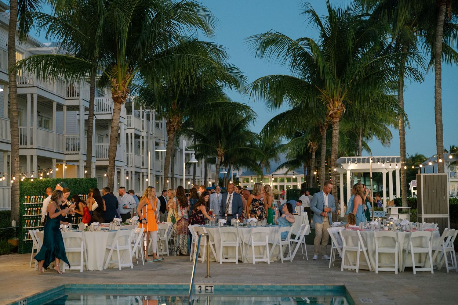 Outdoor beach wedding reception setup with palm trees and white tables at dusk.