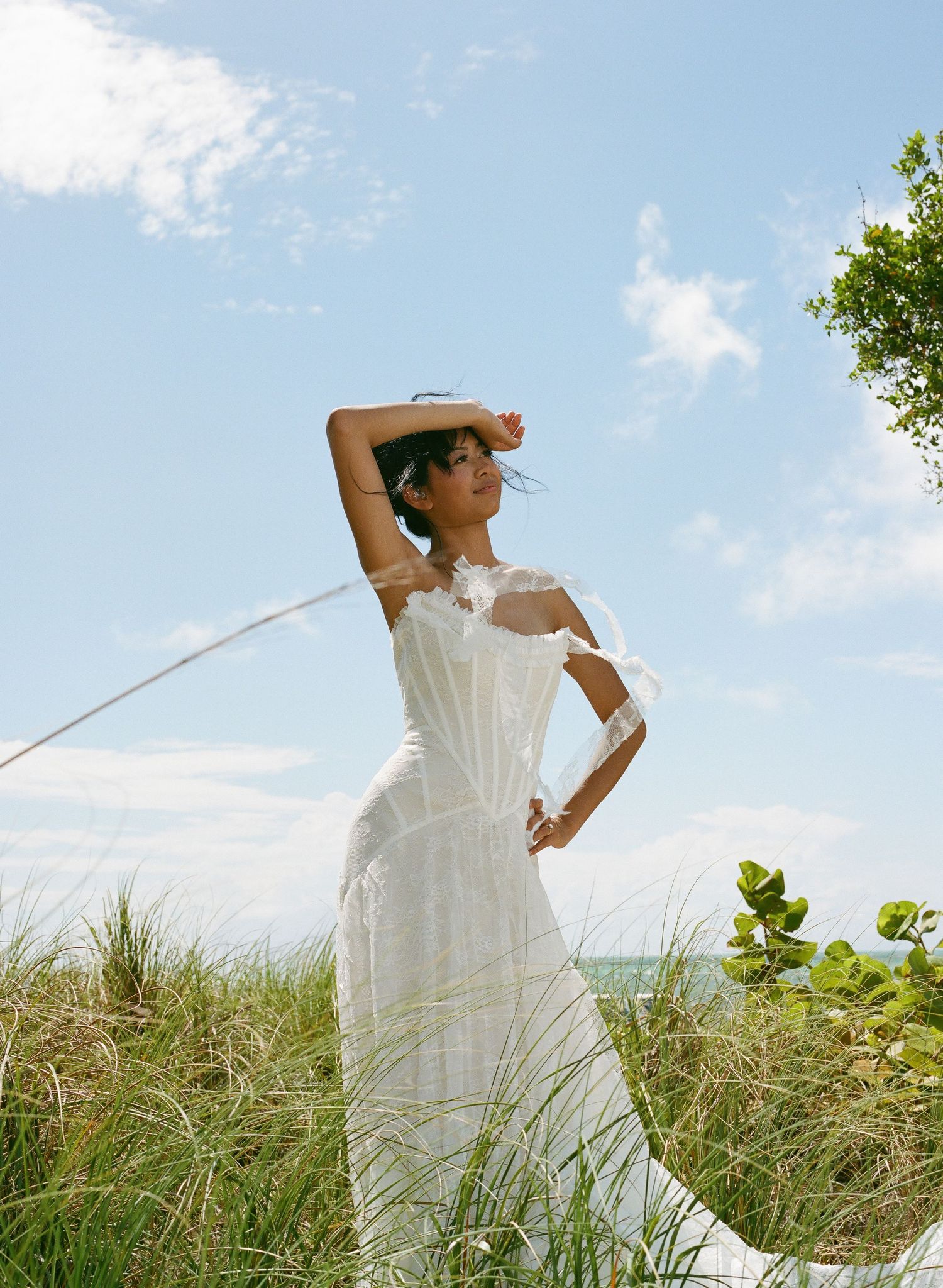 Woman in white off-shoulder maxi dress poses gracefully among coastal dune grass under a bright blue sky.