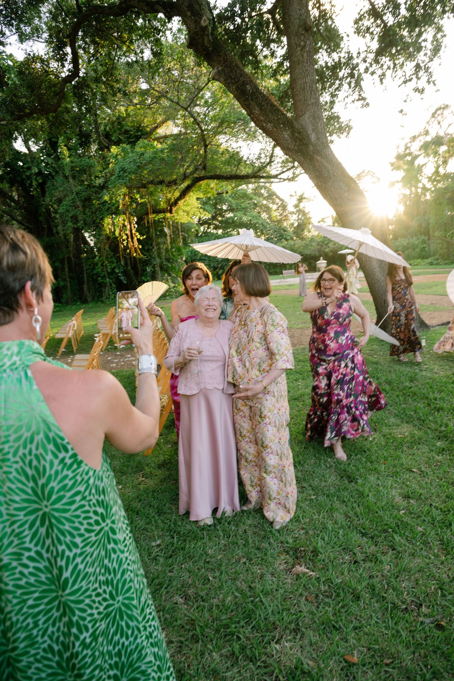 Elegantly dressed wedding guests celebrate outdoors at golden hour, holding parasols and fans beneath large oak trees.