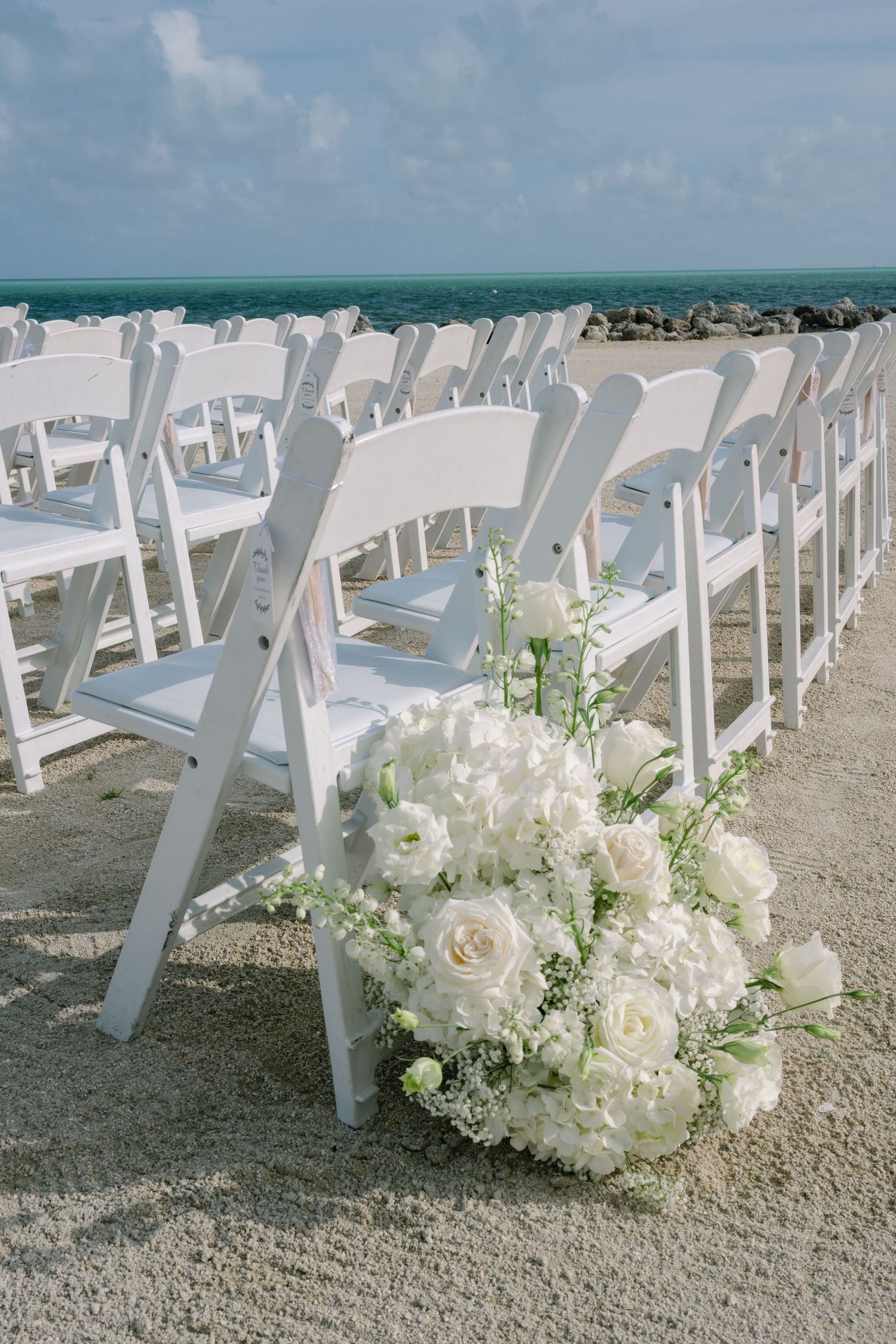 White folding chairs arranged on sandy beach with white floral arrangements for an outdoor wedding ceremony by the ocean.