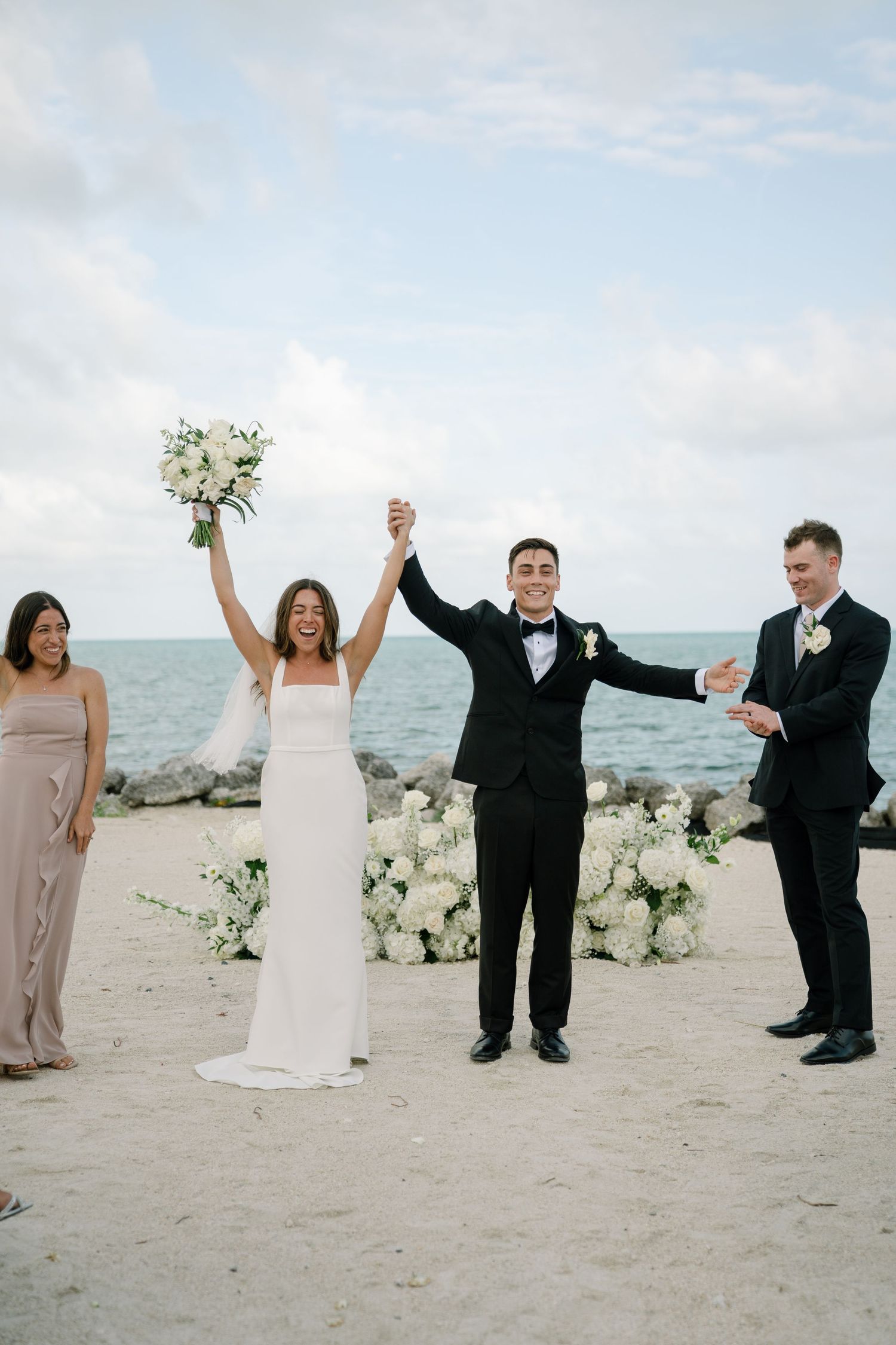 Joyful bride and groom raise their hands in celebration on a sandy beach after their ceremony.