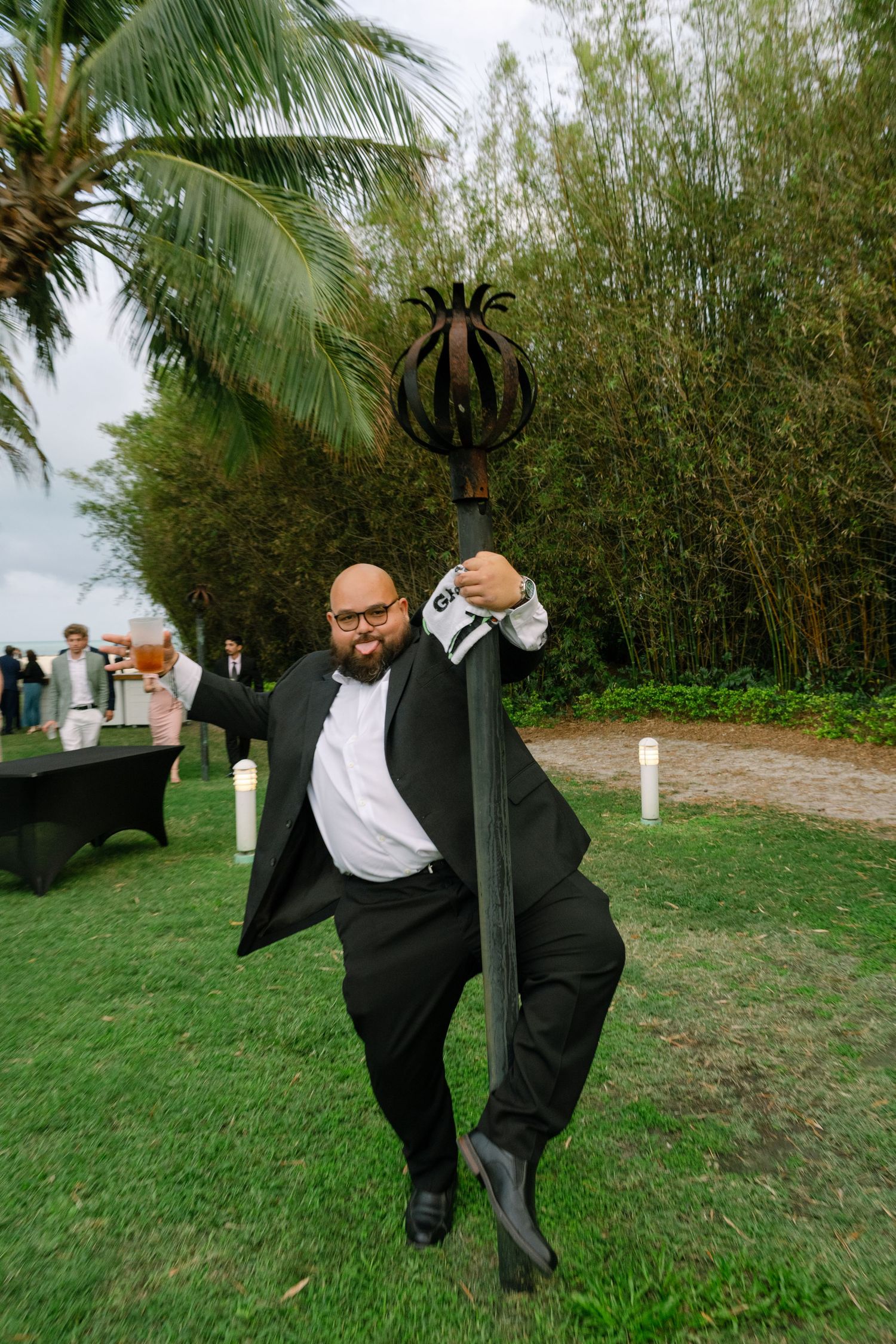 A bald man in a black tuxedo playfully poses with a decorative metal staff on a green lawn at an outdoor wedding.