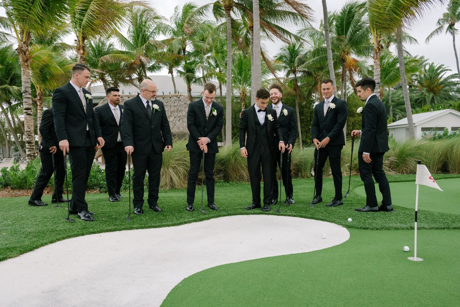 Groomsmen in black suits playing mini golf on putting green surrounded by tropical palm trees.