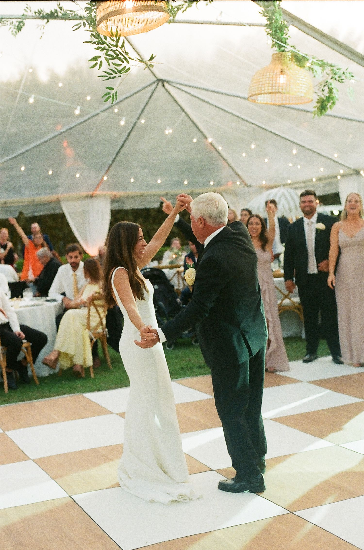 Bride in white gown dancing with father under clear tent with string lights and greenery.