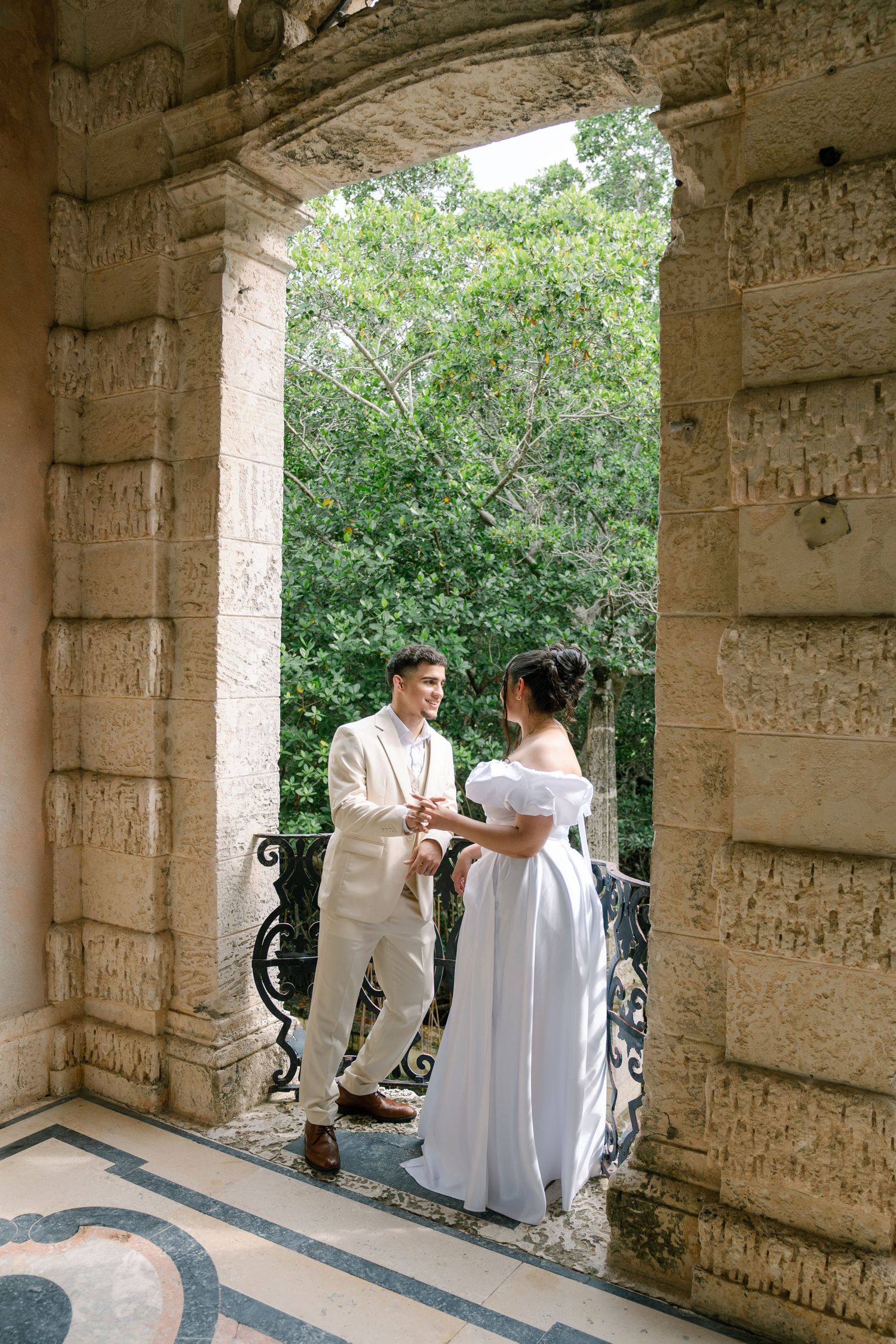 People in white clothing gather under a stone colonnade with classic architectural details and greenery in background.