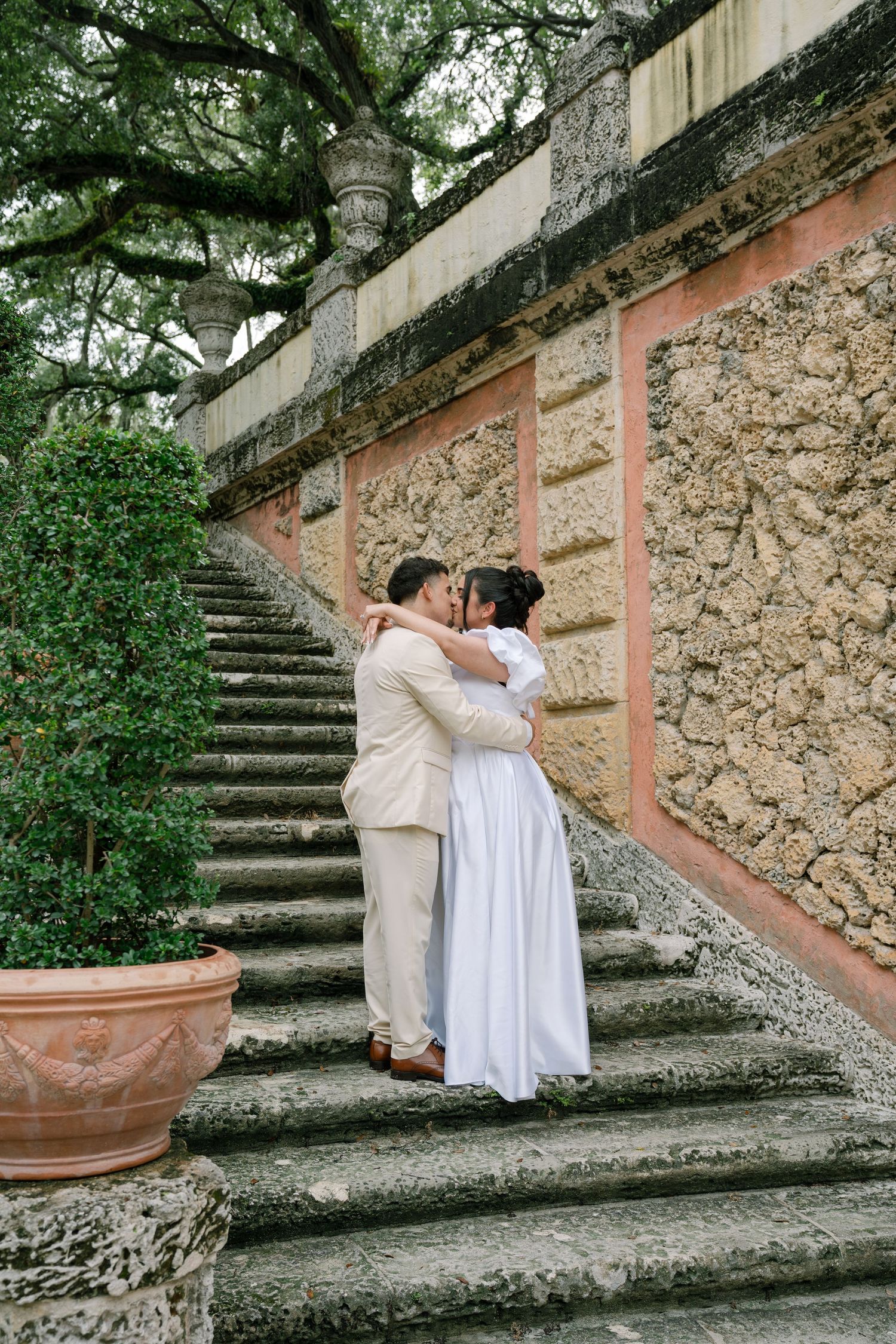 A couple embraces on stone garden steps next to a rustic coral-colored wall and lush greenery at a historic estate.