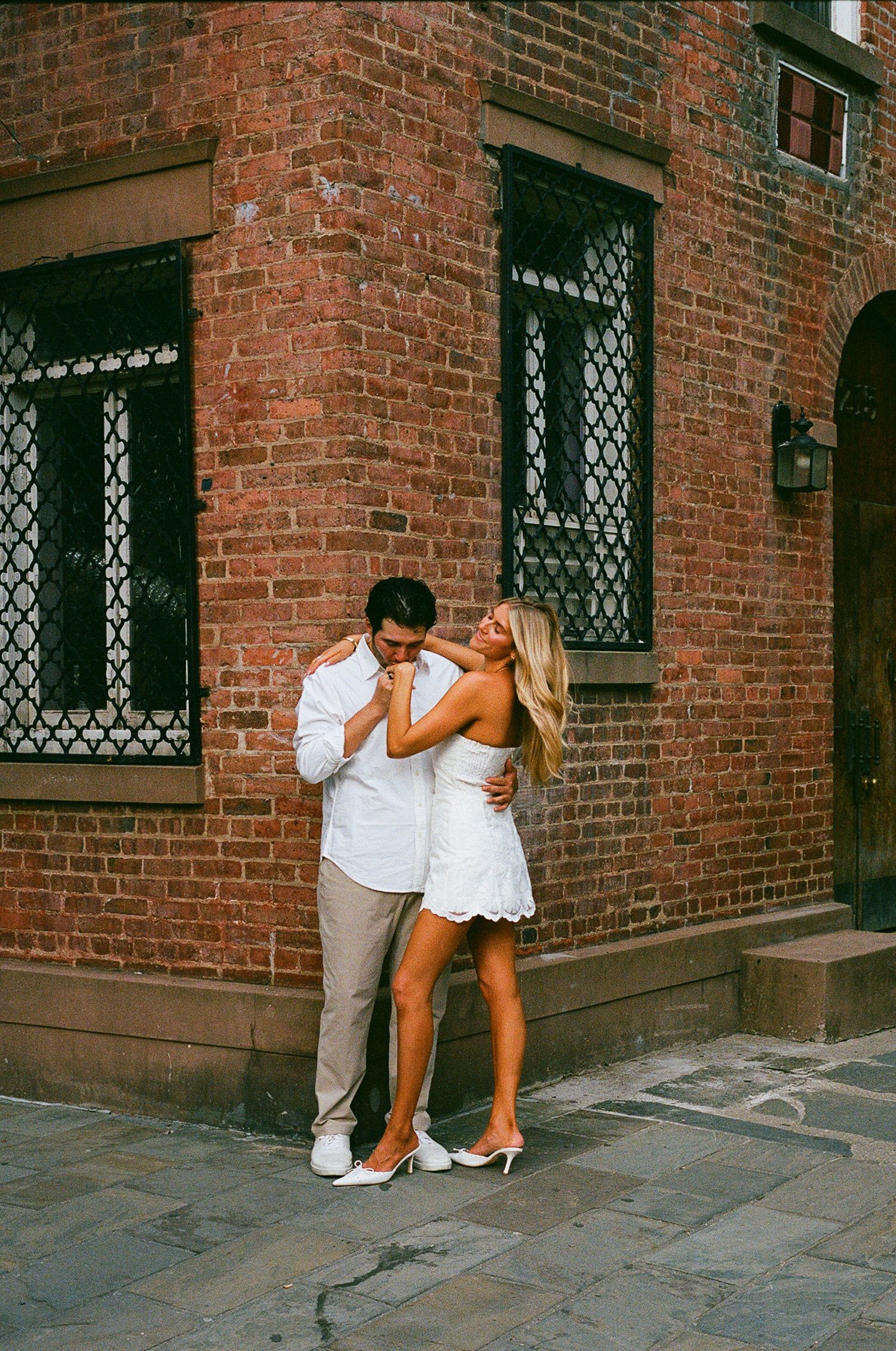 Couple embracing romantically in front of a rustic red brick building, woman in white dress and man in casual attire.