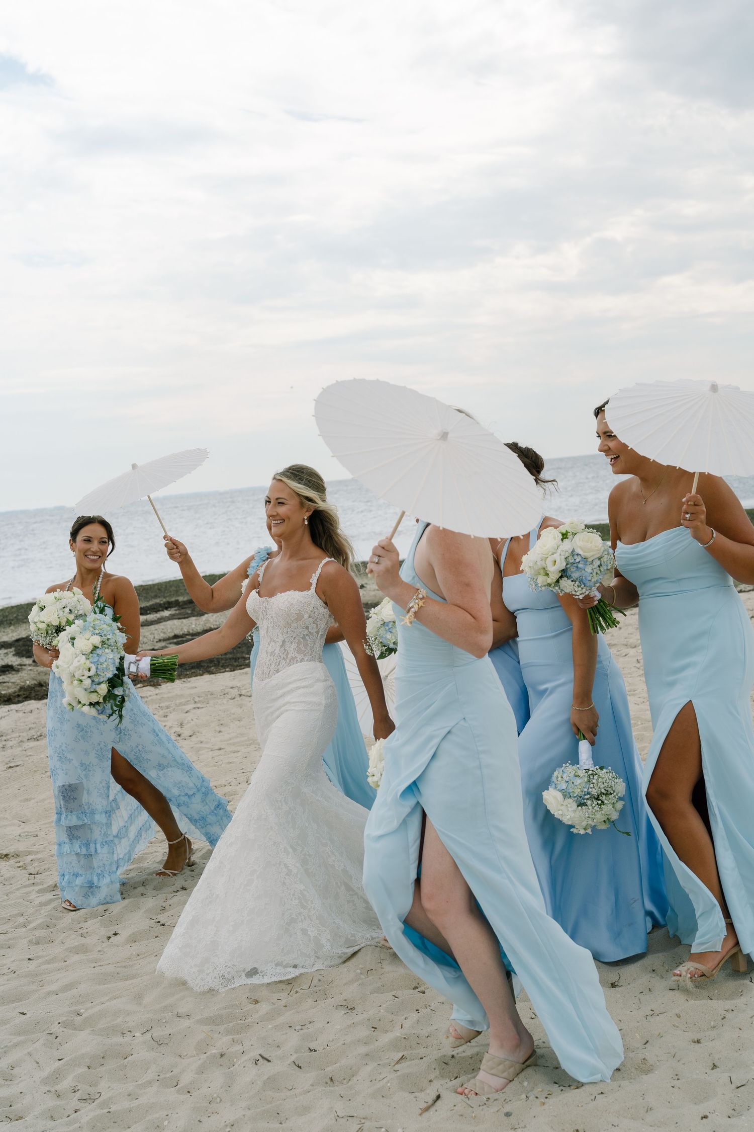 Bride and bridesmaids in light blue dresses walk on beach holding white parasols and bouquets on a cloudy day.