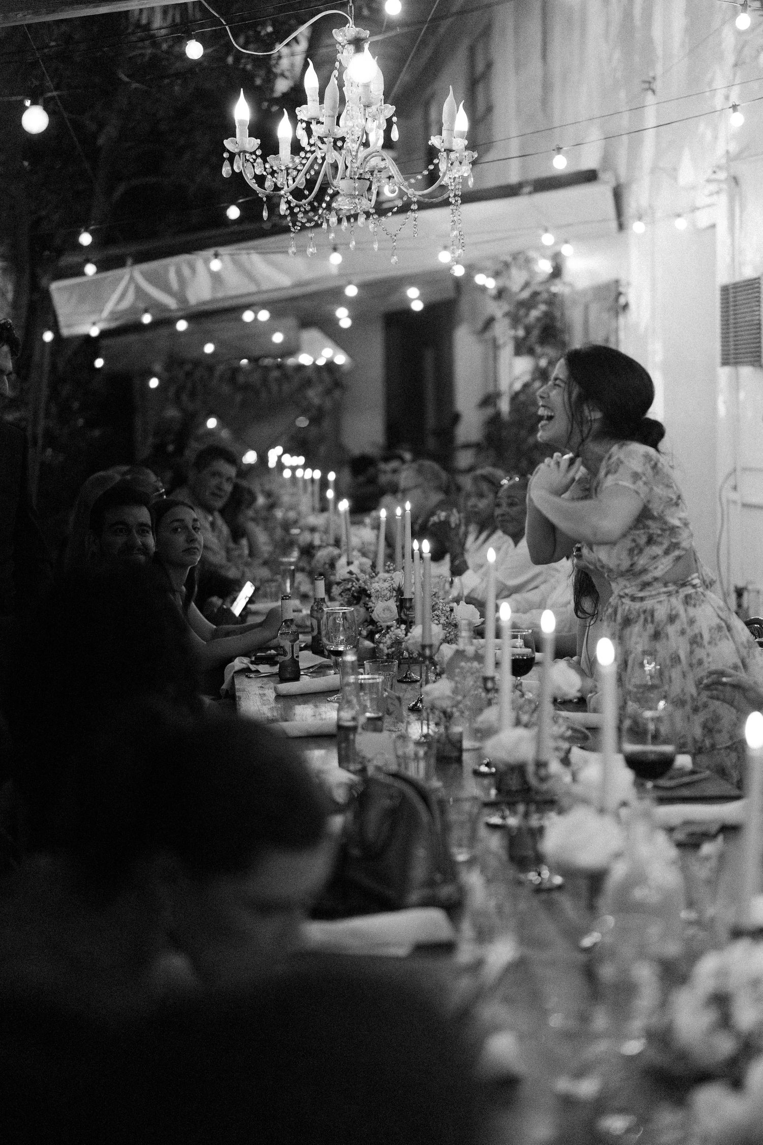 Guests laugh during candlelit wedding reception dinner under chandelier and string lights in black and white photo.
