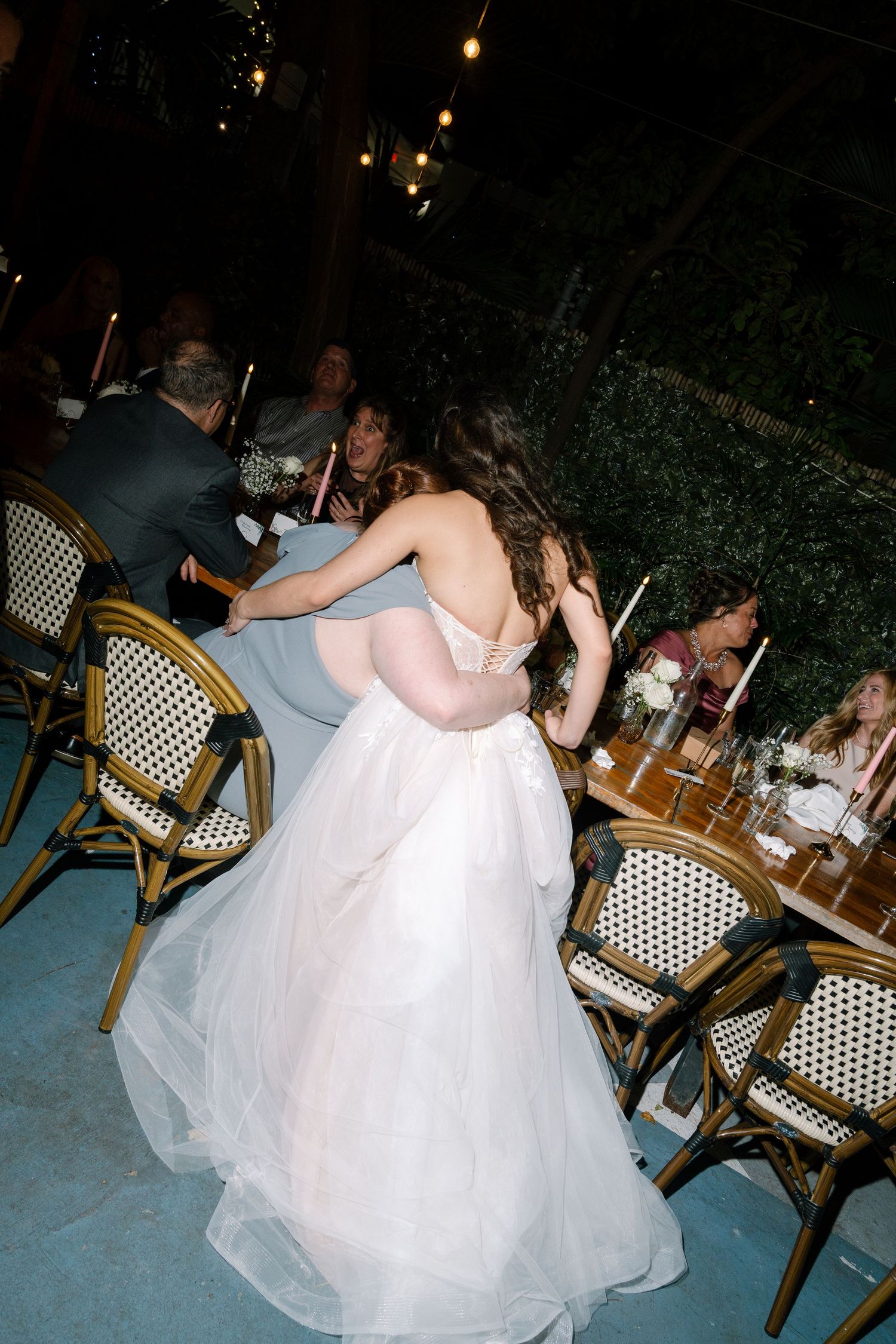 Bride in elegant backless tulle ballgown embracing guest at outdoor wedding reception with string lights.