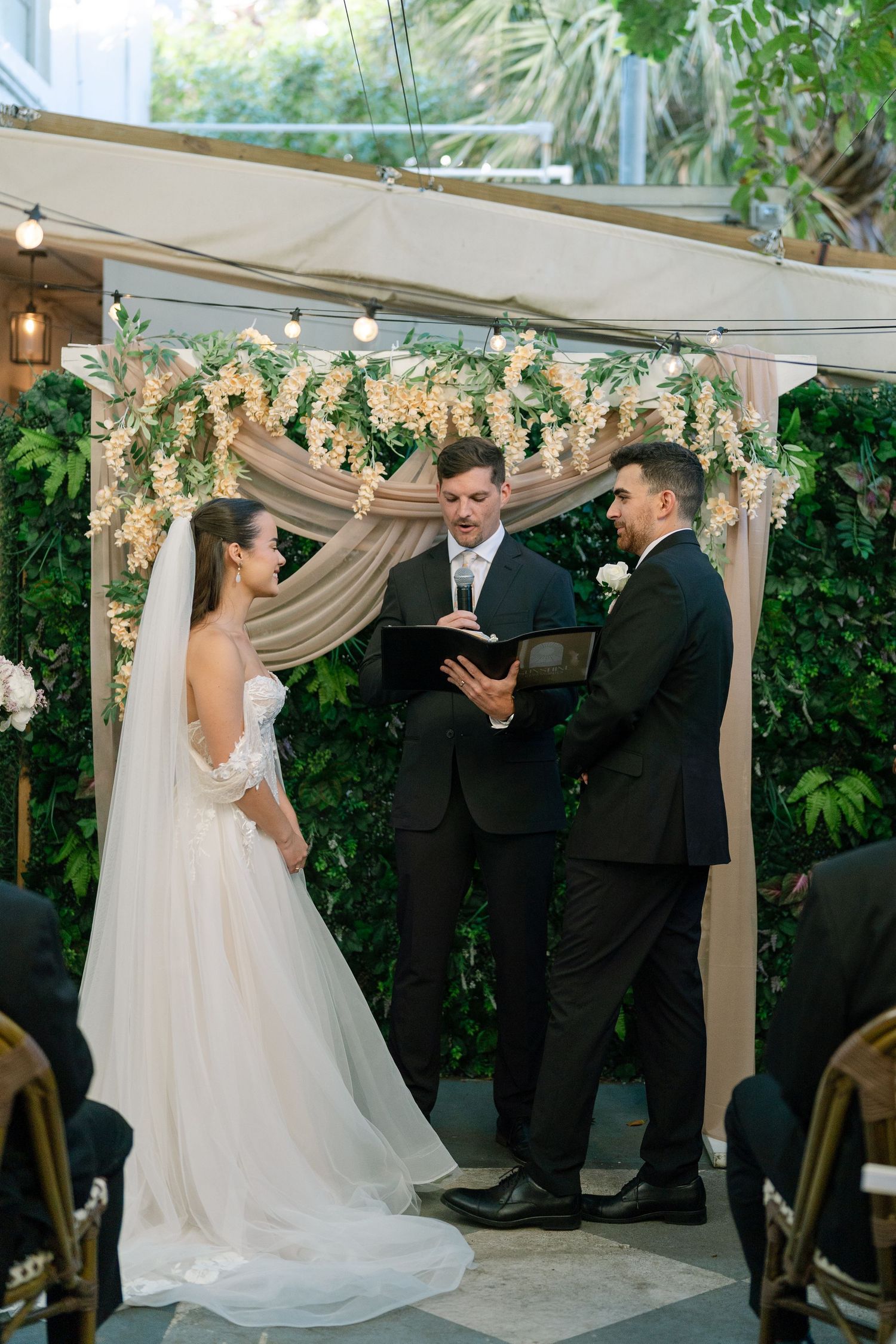 Bride and groom exchange vows under floral arch with officiant in elegant indoor wedding ceremony.