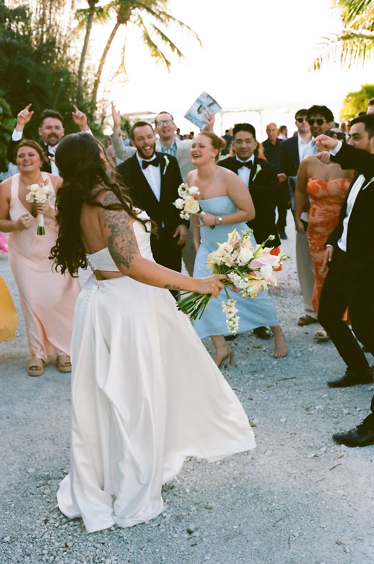 Bride in white gown holds bouquet while guests celebrate at an outdoor tropical wedding reception.