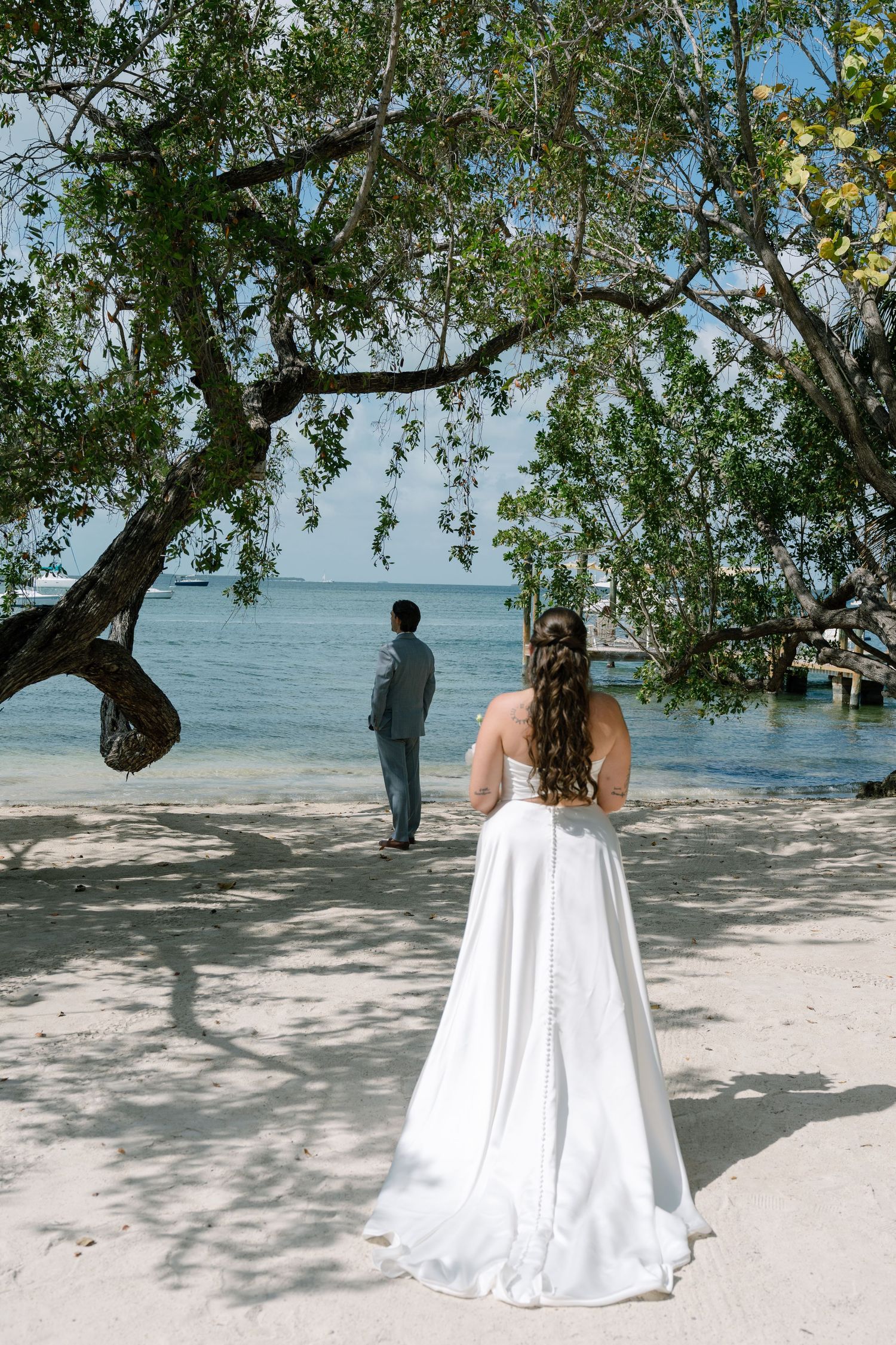Bride in white gown stands on sandy beach facing groom by turquoise water, framed by tropical tree branches.