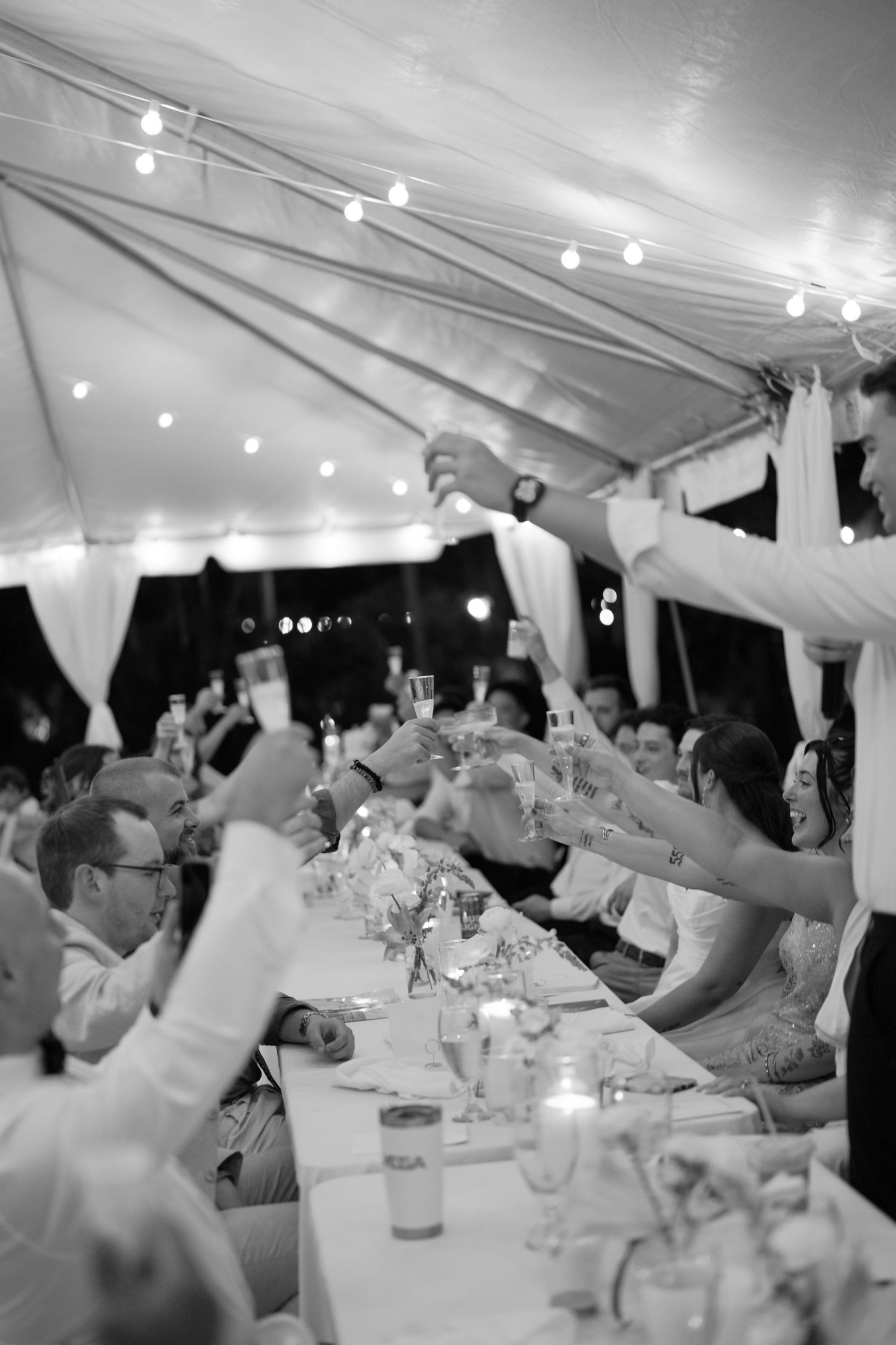 Black and white photo of wedding guests raising glasses in a toast under a tent with string lights.