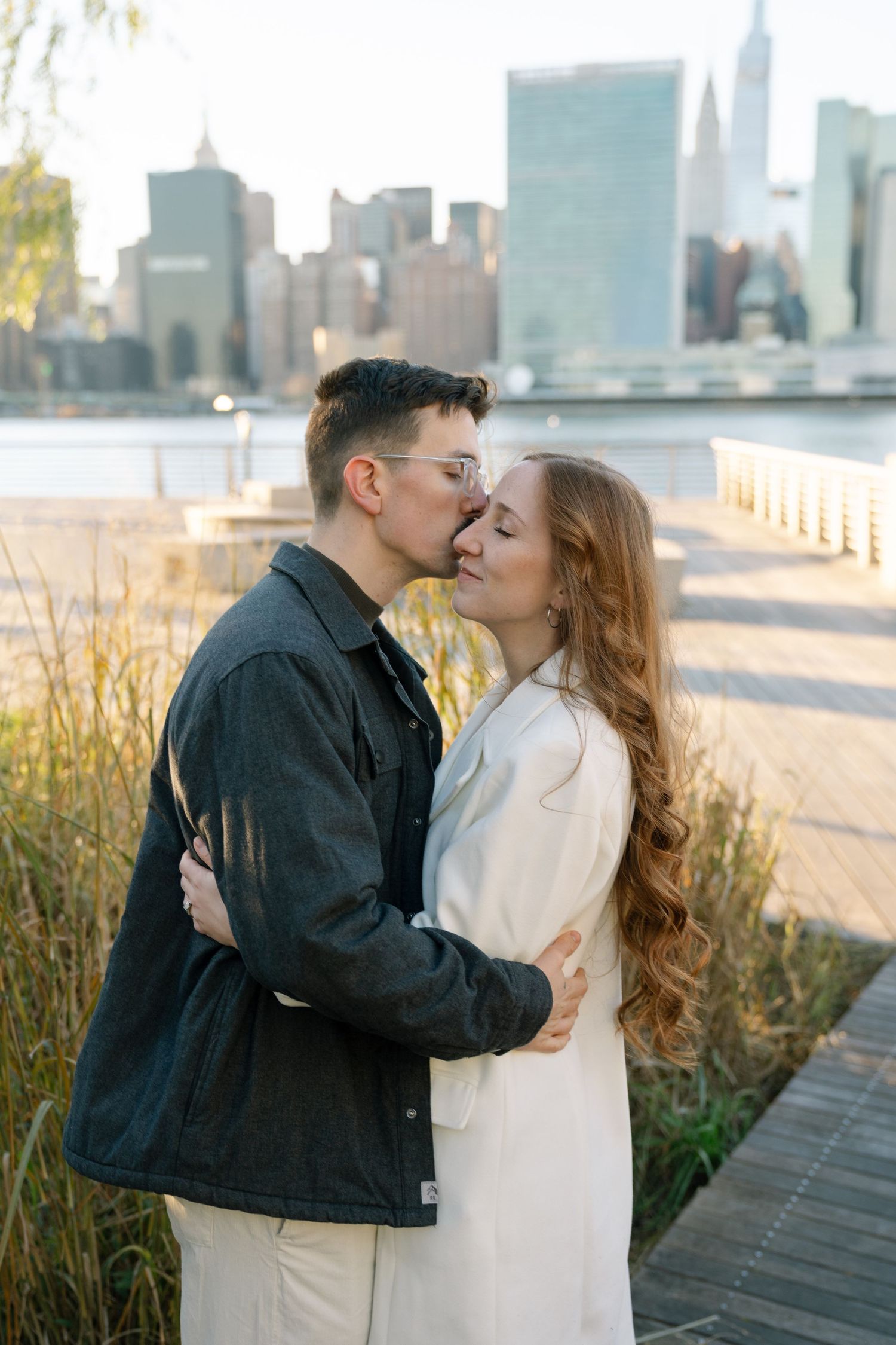 Intimate moment captured on wooden boardwalk with Manhattan skyline and natural grasses.