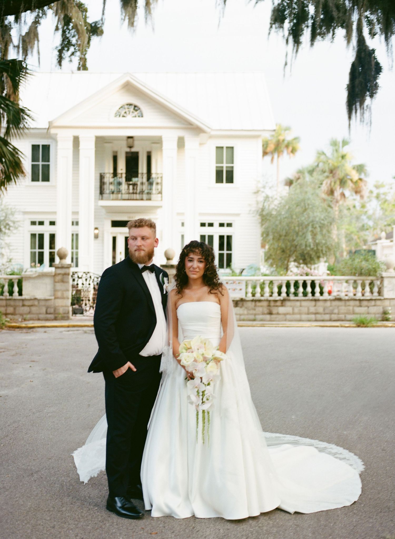 Elegant wedding couple poses in front of a grand white Southern mansion with Spanish moss trees.