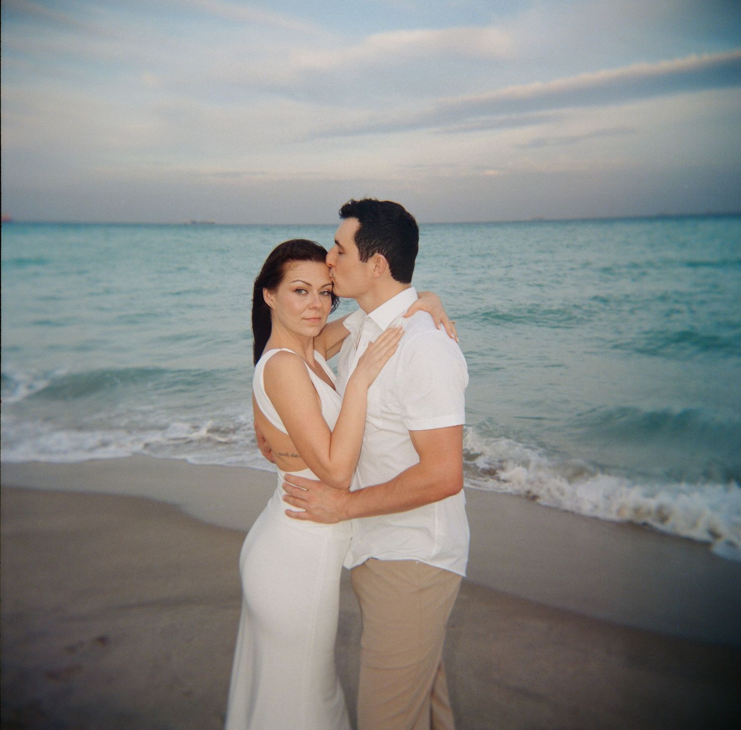 Romantic beach photoshoot of a couple in white and khaki embracing by the ocean at dusk.