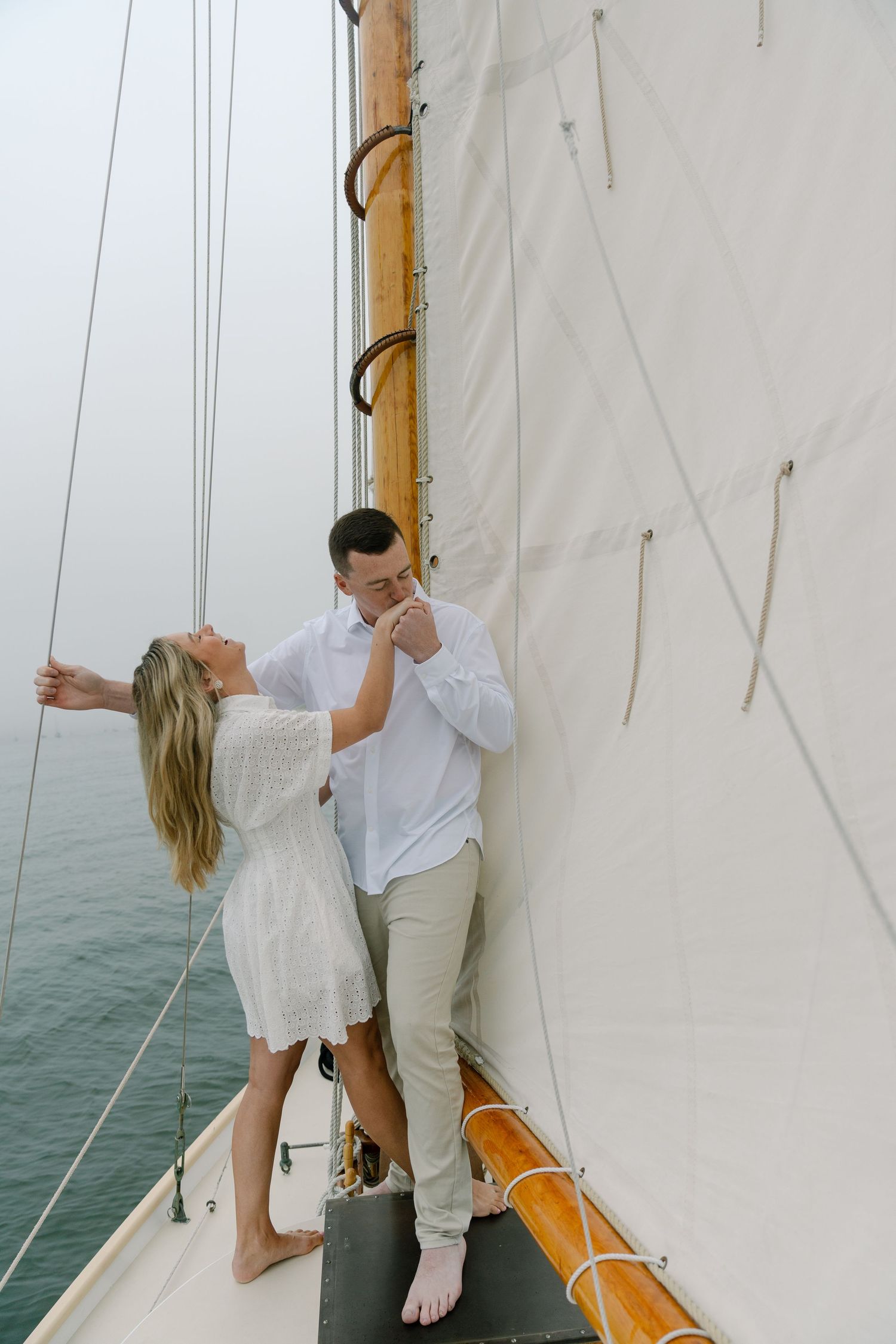 Couple sharing a romantic moment on a sailboat deck, dressed in white, with ocean views and billowing sails behind them.