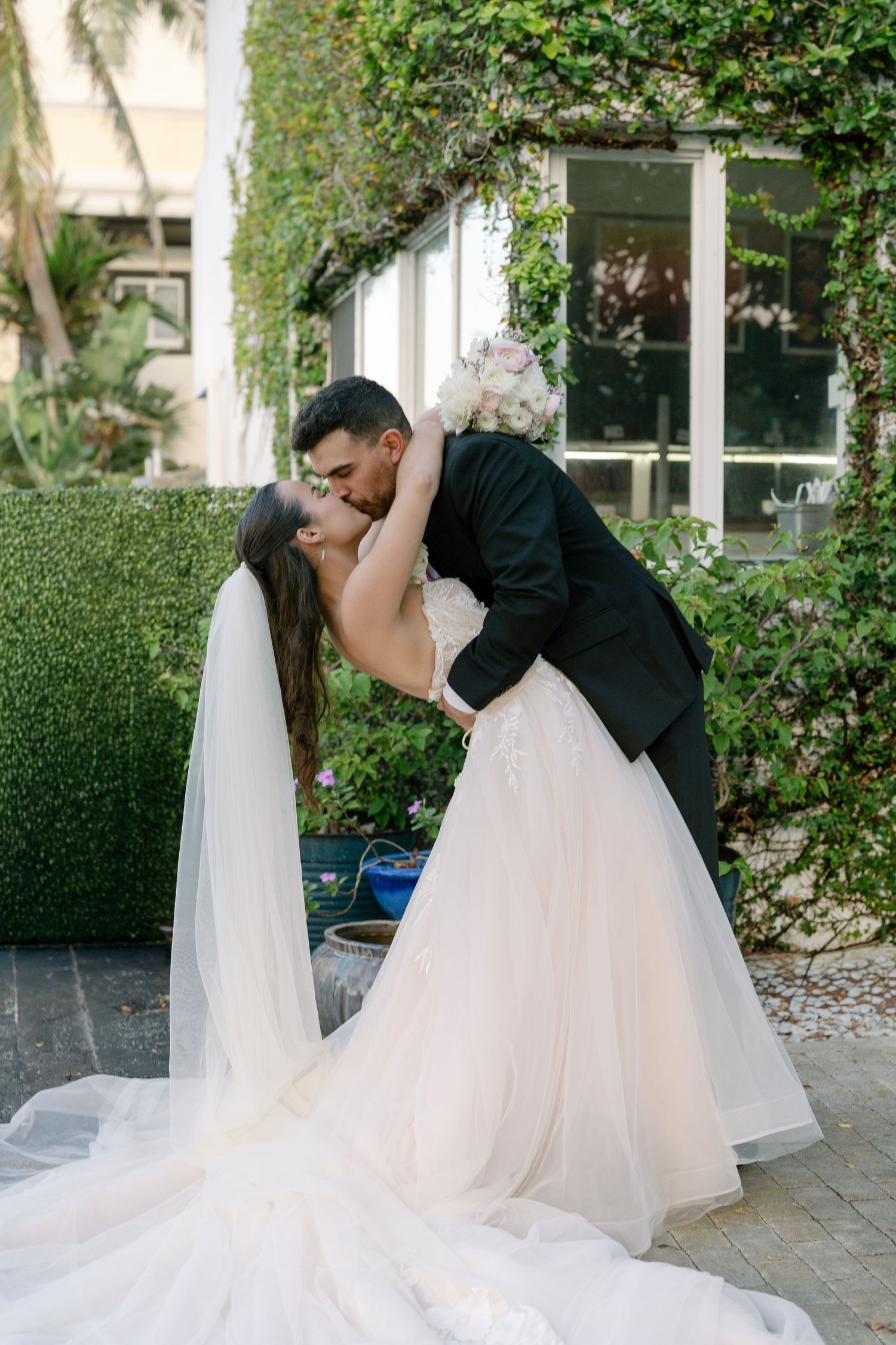 Bride and groom share a romantic dip kiss outdoors, surrounded by lush greenery, as she holds a white bouquet.