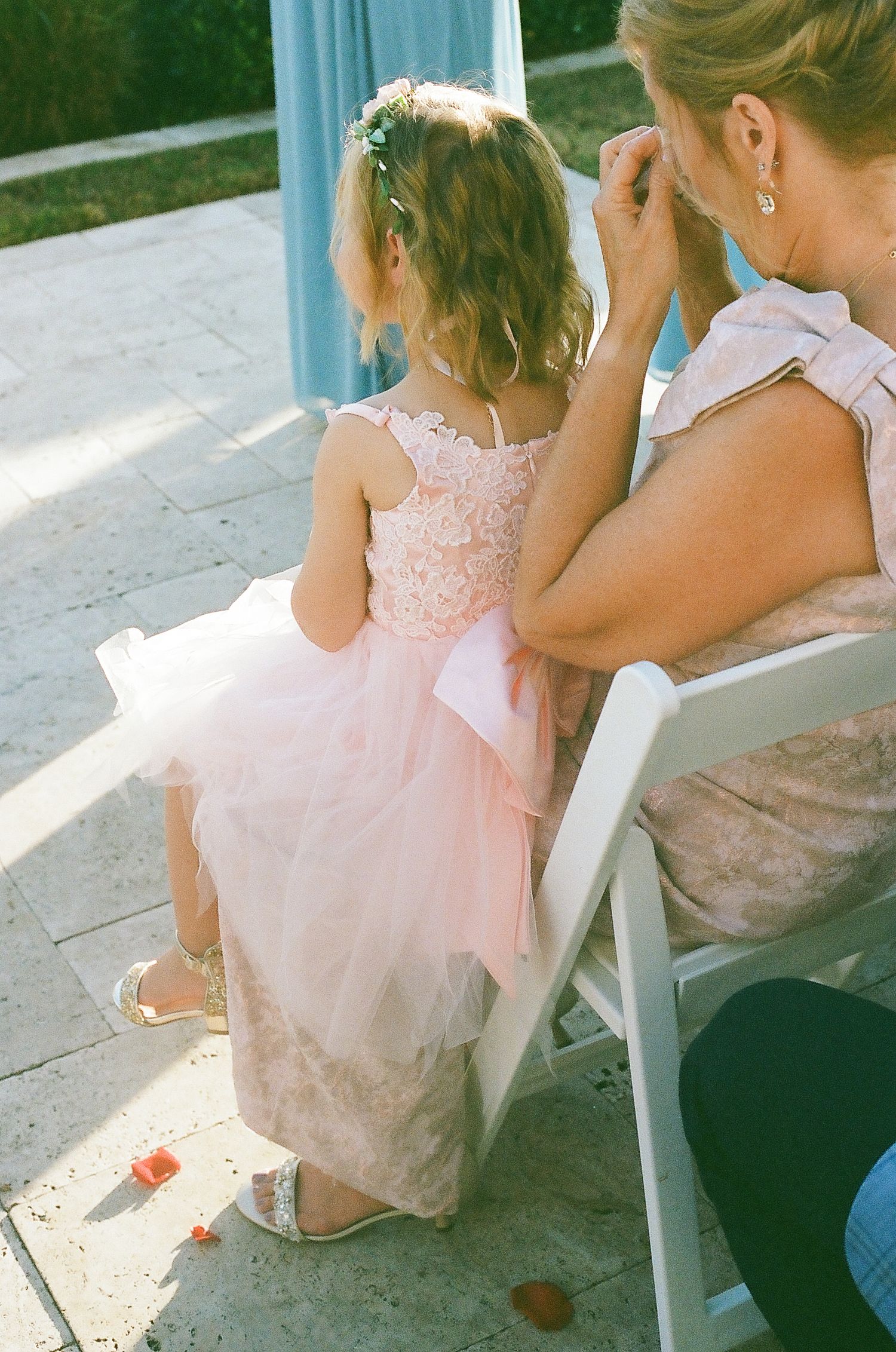 A young girl in a pink tulle dress sits on a white chair during a sunny outdoor event.